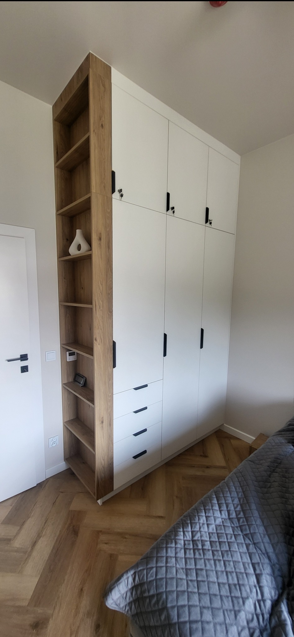 Wysoka, biała szafa z czterema górnymi drzwiczkami zamykanymi na klucz, trzema szufladami i otwartym regałem z drewnianymi półkami, uzupełniona wazonem i elektronicznym zegarkiem, na parkiecie...