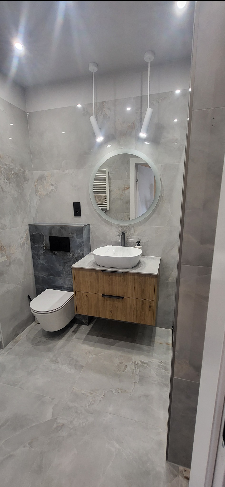 Nowoczesna łazienka z umywalką nablatową, szafką z drewnopodobnym frontem, okrągłym lustrem z podświetleniem LED i wiszącą toaletą, wykończona płytkami imitującymi marmur.