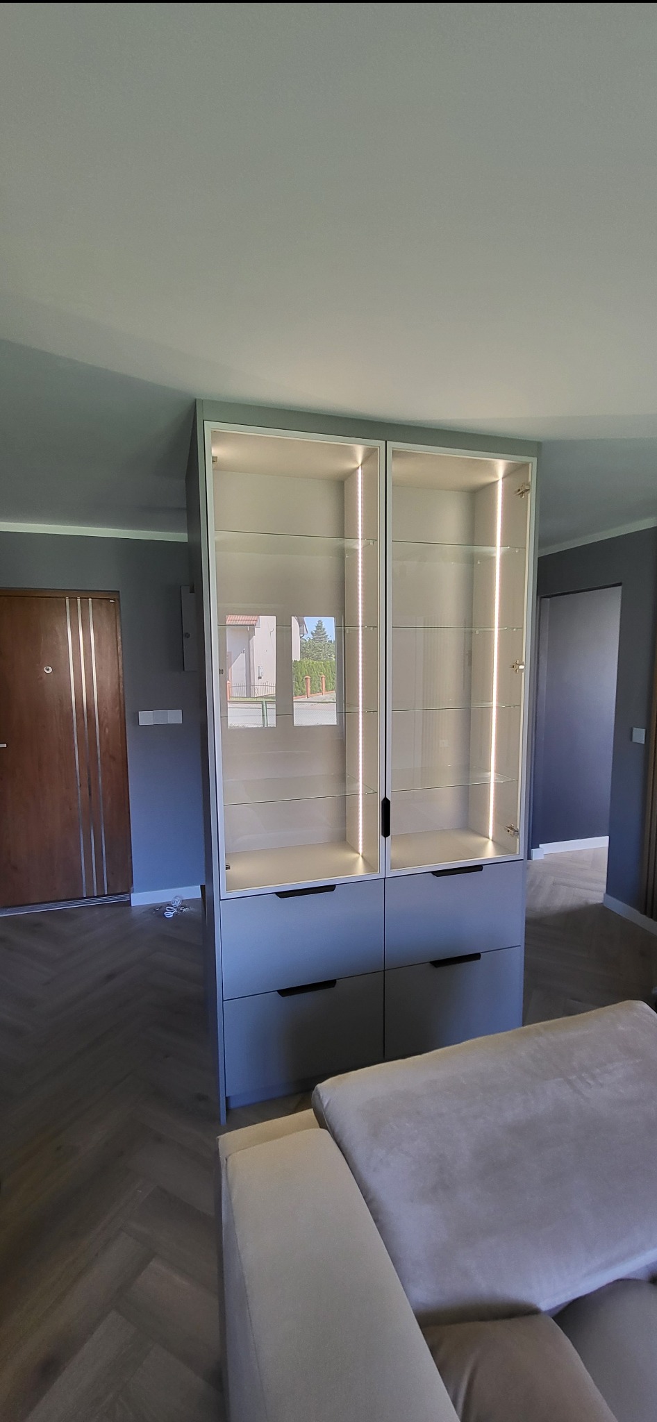 Nowoczesna, szara witryna z przeszklonymi drzwiami i wewnętrznym oświetleniem LED, stojąca w salonie z parkietem w jodełkę i szarą ścianą.