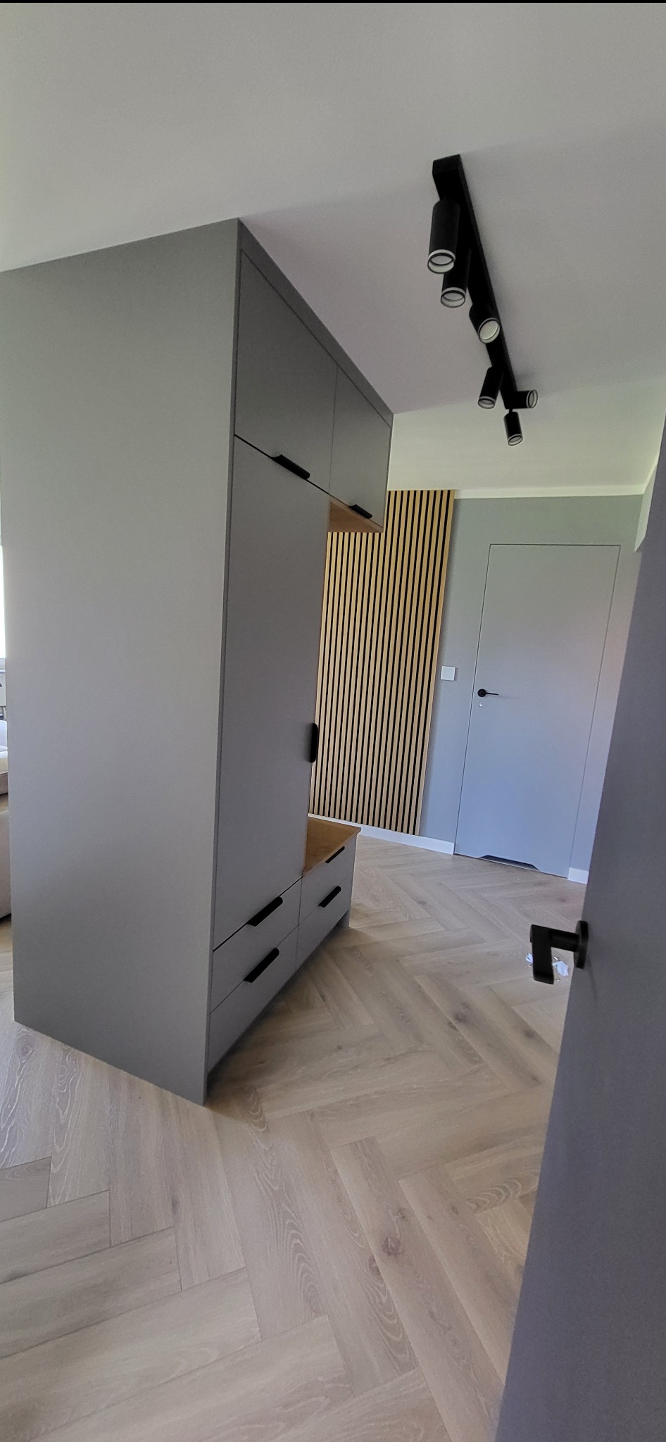 Szara szafa na wymiar z czarnymi uchwytami, drewniana lamela na ścianie w tle, parkiet w jodełkę.