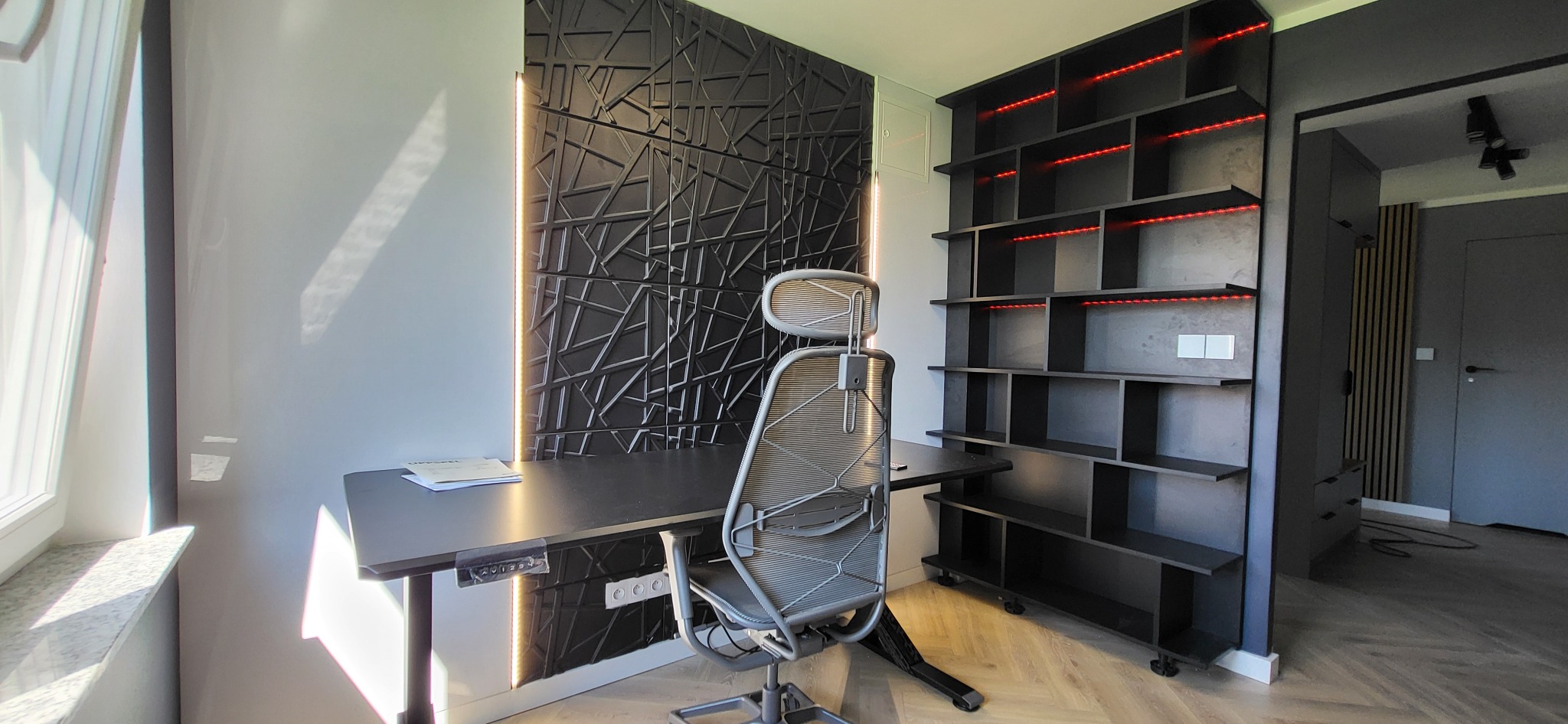 Nowoczesne biuro z czarnym biurkiem z regulowaną wysokością, ergonomicznym krzesłem, modułową szafą regałową z podświetleniem LED i dekoracyjną ścianą 3D z oświetleniem.