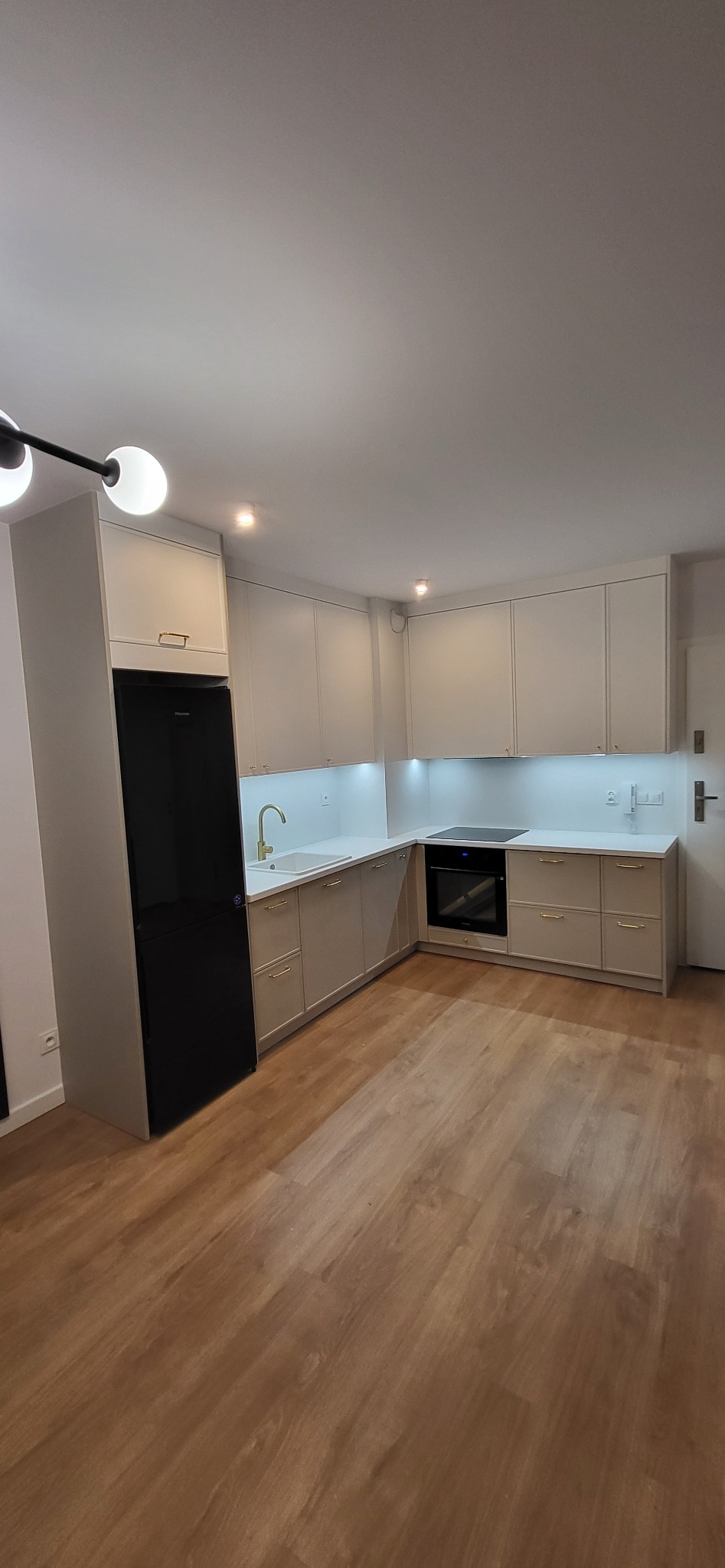 Jasna kuchnia w kształcie litery L z frontami bez uchwytów, złotą armaturą i czarną lodówką, oświetlona punktowymi lampami i listwą LED pod szafkami wiszącymi.