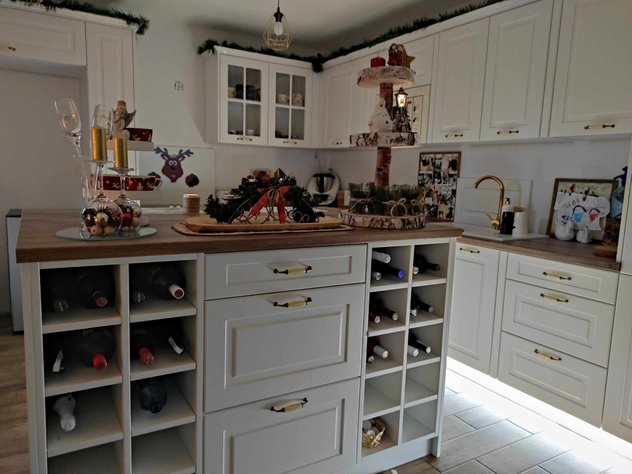 Jasna kuchnia na wymiar z drewnianym blatem i wbudowanym regałem na wino, udekorowana świątecznie zielonymi girlandami i ozdobami.
