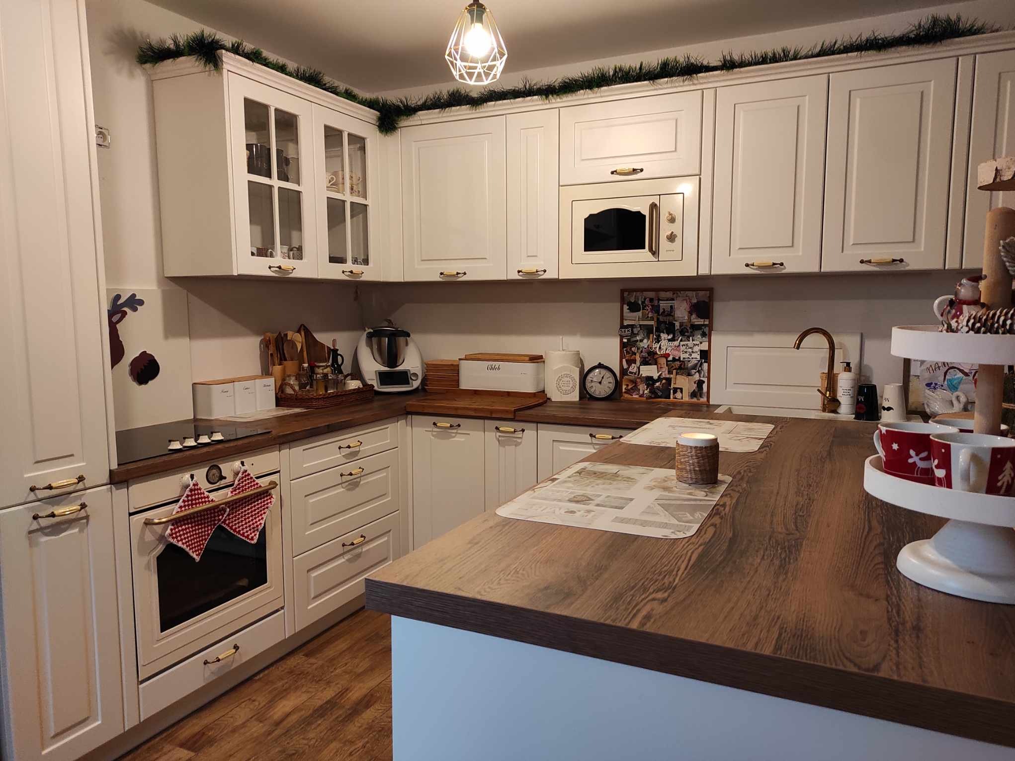 Biała kuchnia z drewnianymi blatami i złotymi uchwytami, ozdobiona świątecznymi girlandami i akcentami, widok na piekarnik z zawieszonymi ręcznikami w czerwoną kratę.