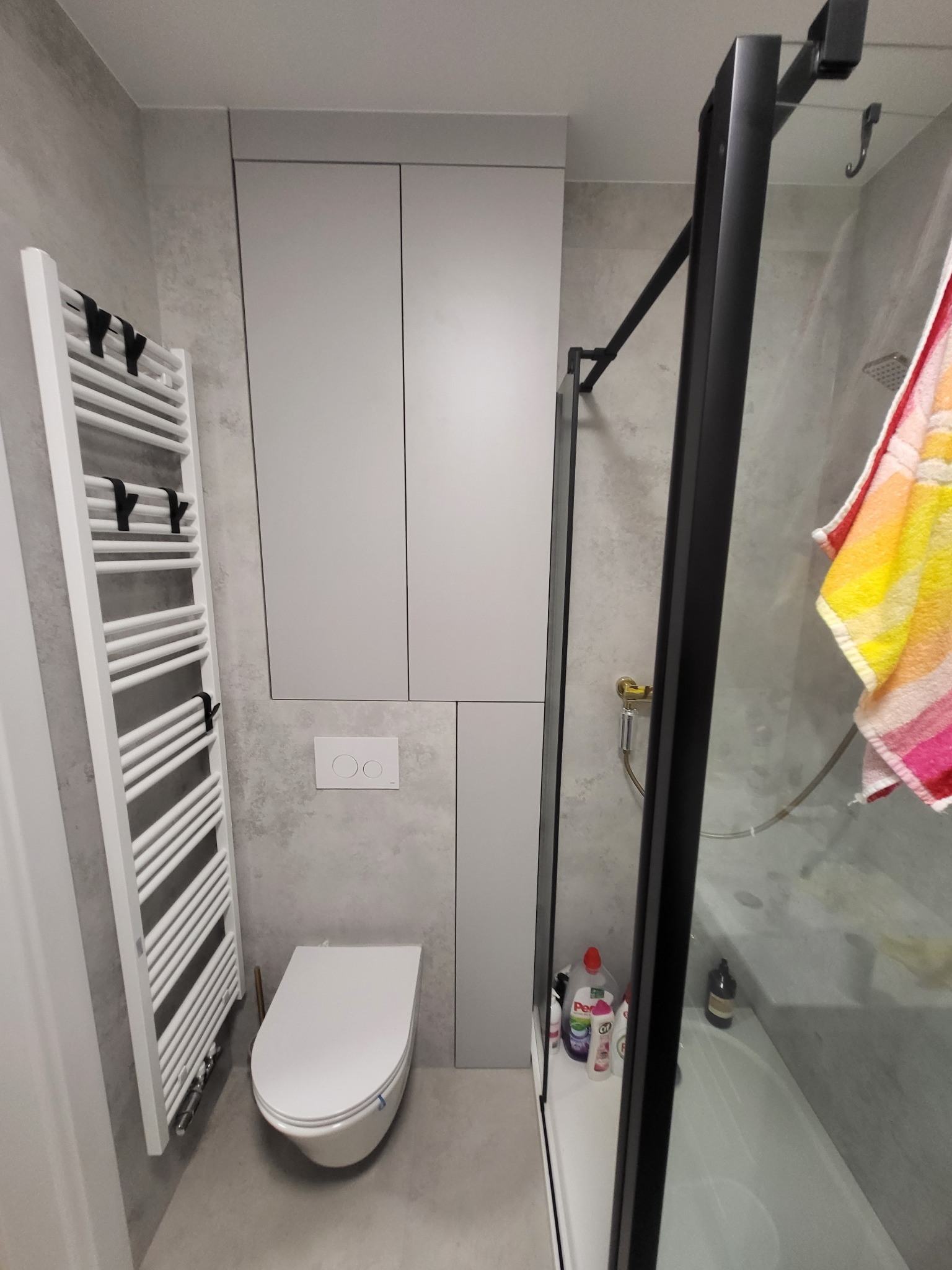 Nowoczesna łazienka z szarą okładziną, wiszącą toaletą, grzejnikiem drabinkowym i kabiną prysznicową z czarną ramą.