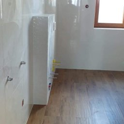 Mast-rem - Nowa łazienka z jasnymi, błyszczącymi płytkami na ścianach i panelami podłogowymi imitującymi drewno, widoczny stelaż podtynkowy WC z wystającymi rurami.