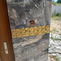 Mast-rem - Fragment ściany wyłożonej płytkami imitującymi marmur z dekoracyjnym złotym pasem z geometrycznym wzorem, widoczne gniazdo elektryczne i stos potłuczonych płytek w tle.