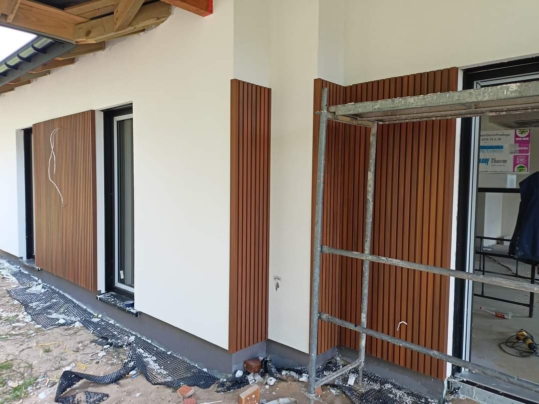 Montaż paneli dekoracyjnych z drewna na elewacji domu jednorodzinnego, widoczne rusztowanie i materiały budowlane.