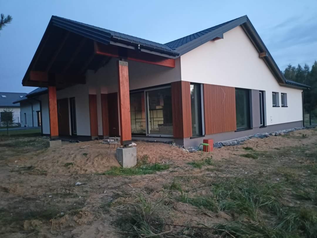 Nowoczesny dom jednorodzinny z drewnianą elewacją i dużymi oknami, widok z zewnątrz na plac budowy przed domem.