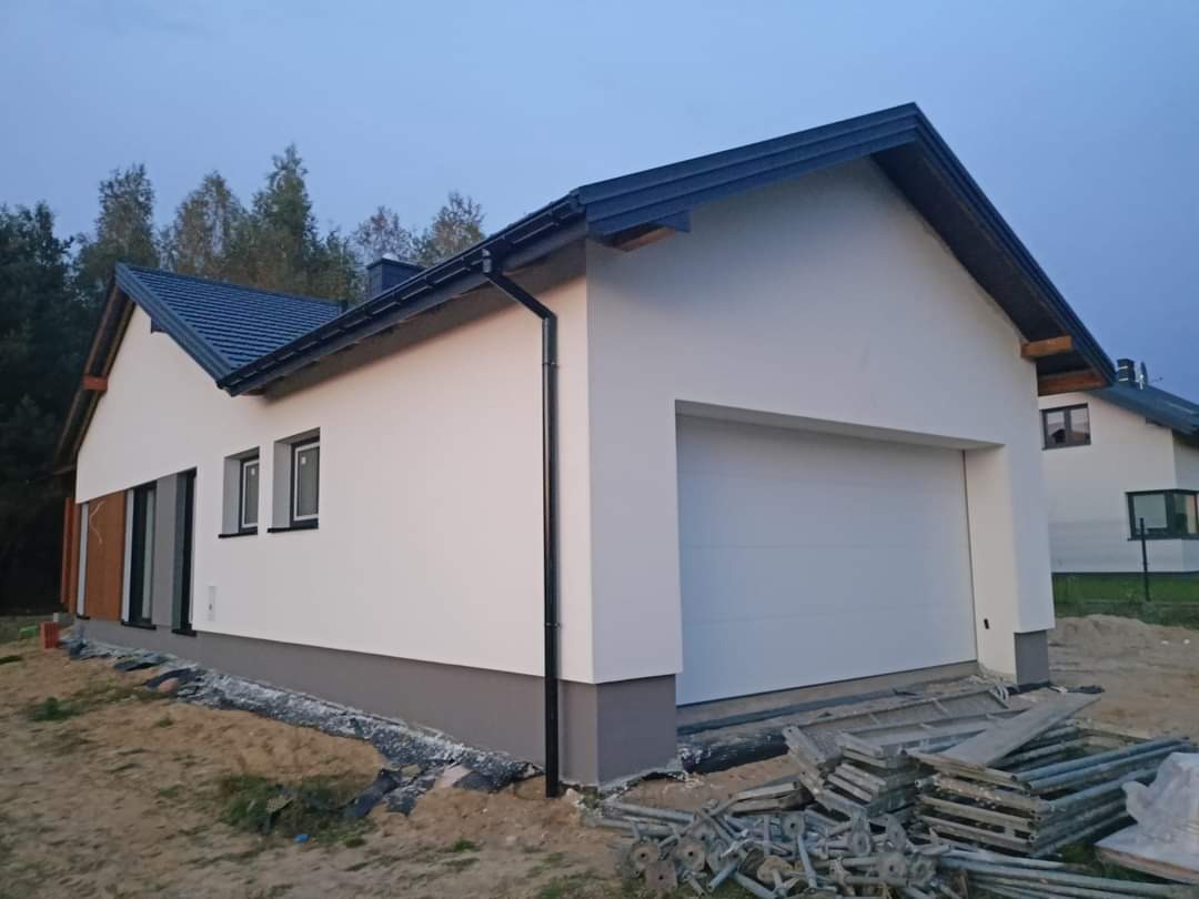 Świeżo otynkowany, parterowy dom jednorodzinny z garażem i ciemną dachówką, widok z boku, z widocznymi rusztowaniami w tle.