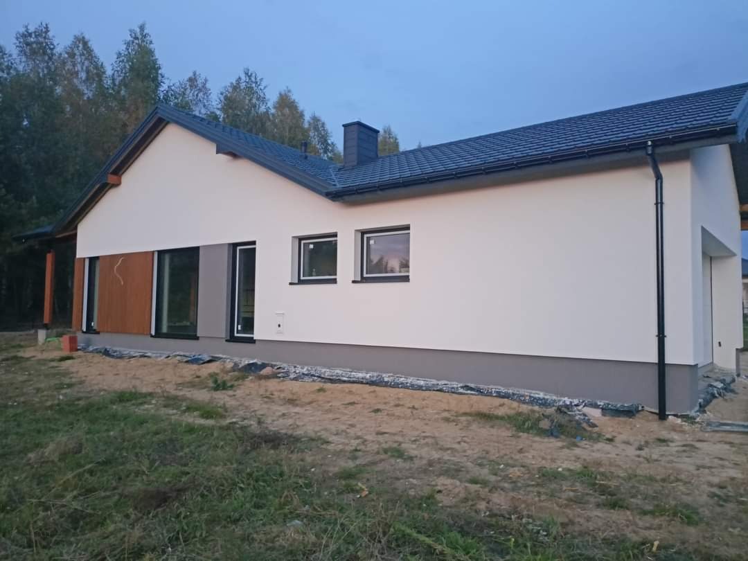 Nowoczesny dom jednorodzinny z białymi ścianami i antracytową stolarką okienną, częściowo wykończony drewnianą okładziną, widok z zewnątrz w trakcie lub po remoncie, na niezagospodarowanej działce.