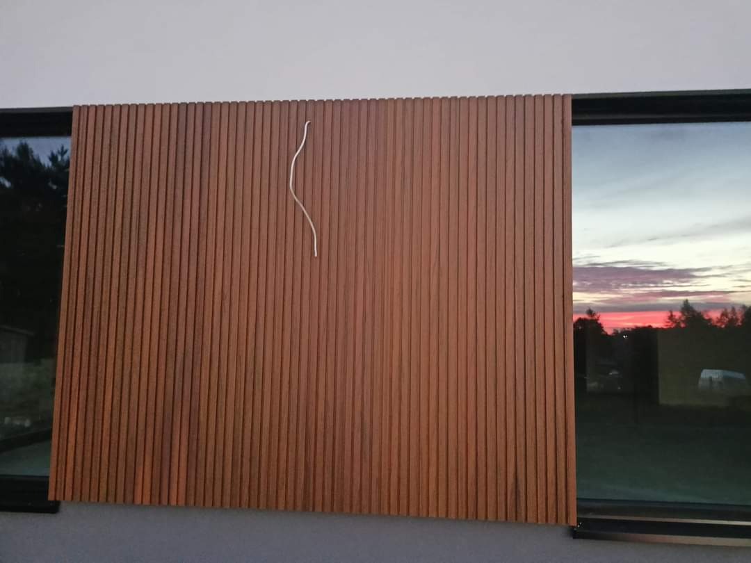 Drewniana okładzina elewacyjna z pionowych listew w kolorze brązu, zamontowana na budynku w Rawie Mazowieckiej, z widocznym srebrnym drutem w kształcie fali i odbiciem zachodu słońca w szybie okna.