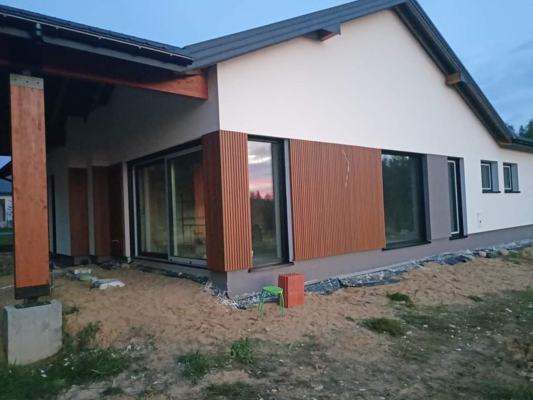 Nowoczesny dom jednorodzinny z drewnianą elewacją w trakcie wykończenia, widoczne elementy konstrukcyjne i przygotowany teren.