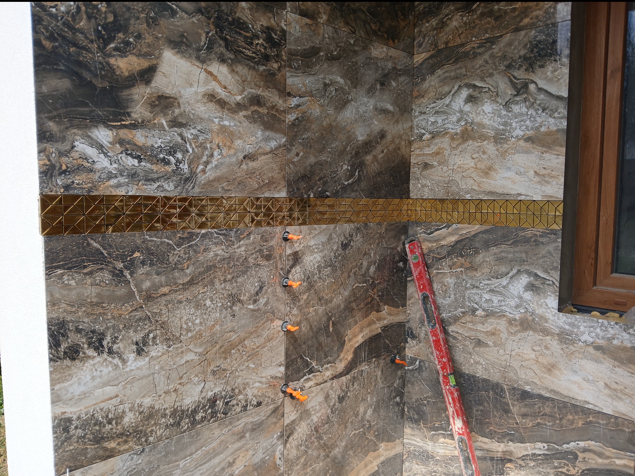 Montaż płytek imitujących marmur z dekoracyjną, złotą listwą, poziomica przy ścianie, widoczne klipsy poziomujące.