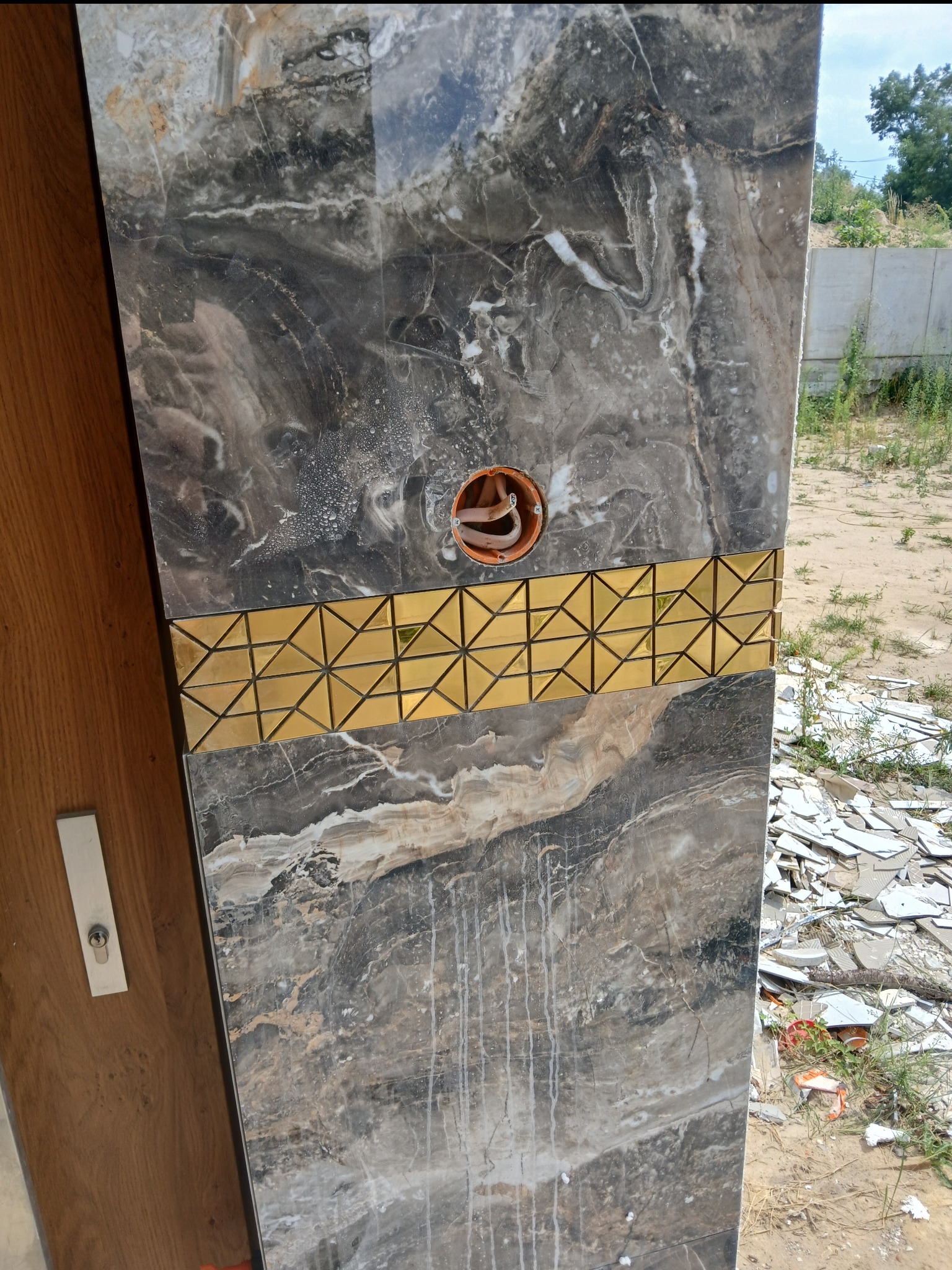 Fragment ściany wyłożonej płytkami imitującymi marmur z dekoracyjnym złotym pasem z geometrycznym wzorem, widoczne gniazdo elektryczne i stos potłuczonych płytek w tle.