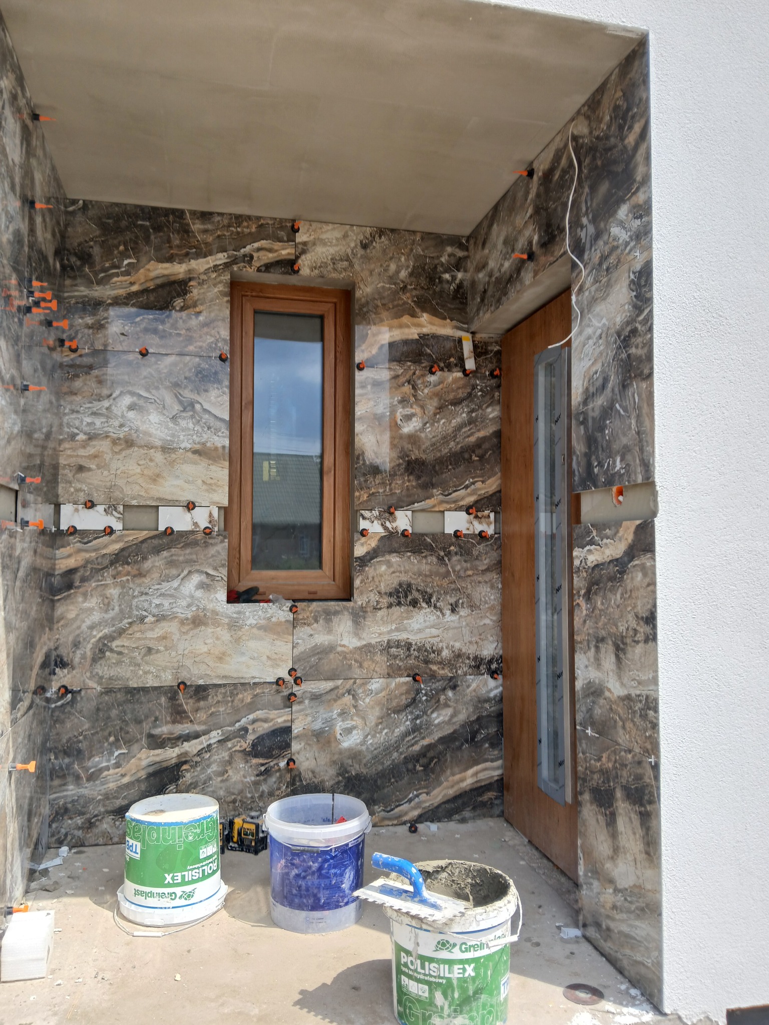 Montaż płytek imitujących marmur w korytarzu wejściowym, widoczne narzędzia glazurnicze i poziomice, pomarańczowe krzyżyki dystansowe, częściowo ułożone płytki wokół okna i drzwi.