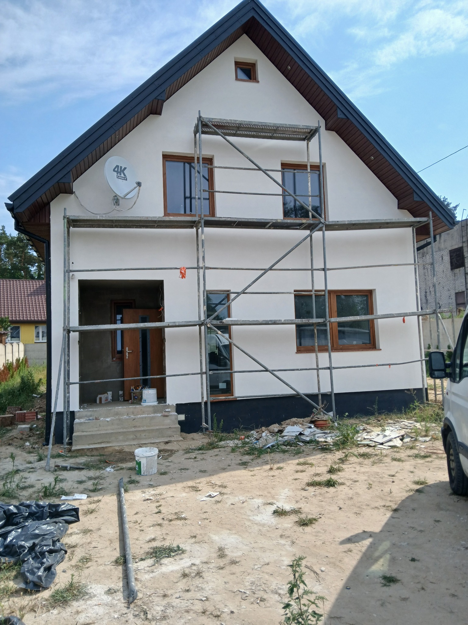 Dom jednorodzinny z białymi ścianami zewnętrznymi w trakcie prac wykończeniowych, widoczne rusztowania, niezabezpieczone wejście i porozrzucane materiały budowlane na podwórku.
