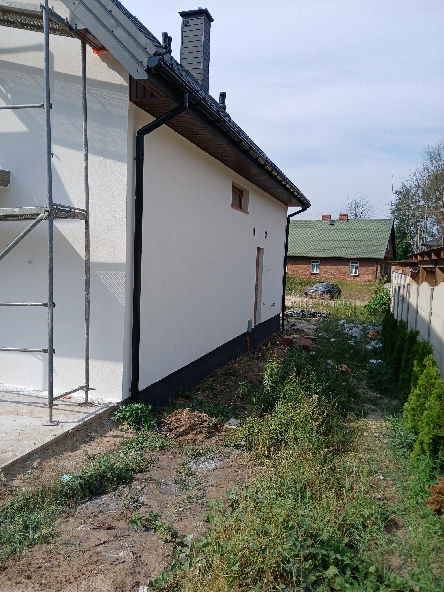 Świeżo otynkowana ściana domu jednorodzinnego z widocznymi rusztowaniami, czarnymi rynnami i fragmentem dachu z ciemną dachówką, na tle sąsiedniego domu z zielonym dachem.