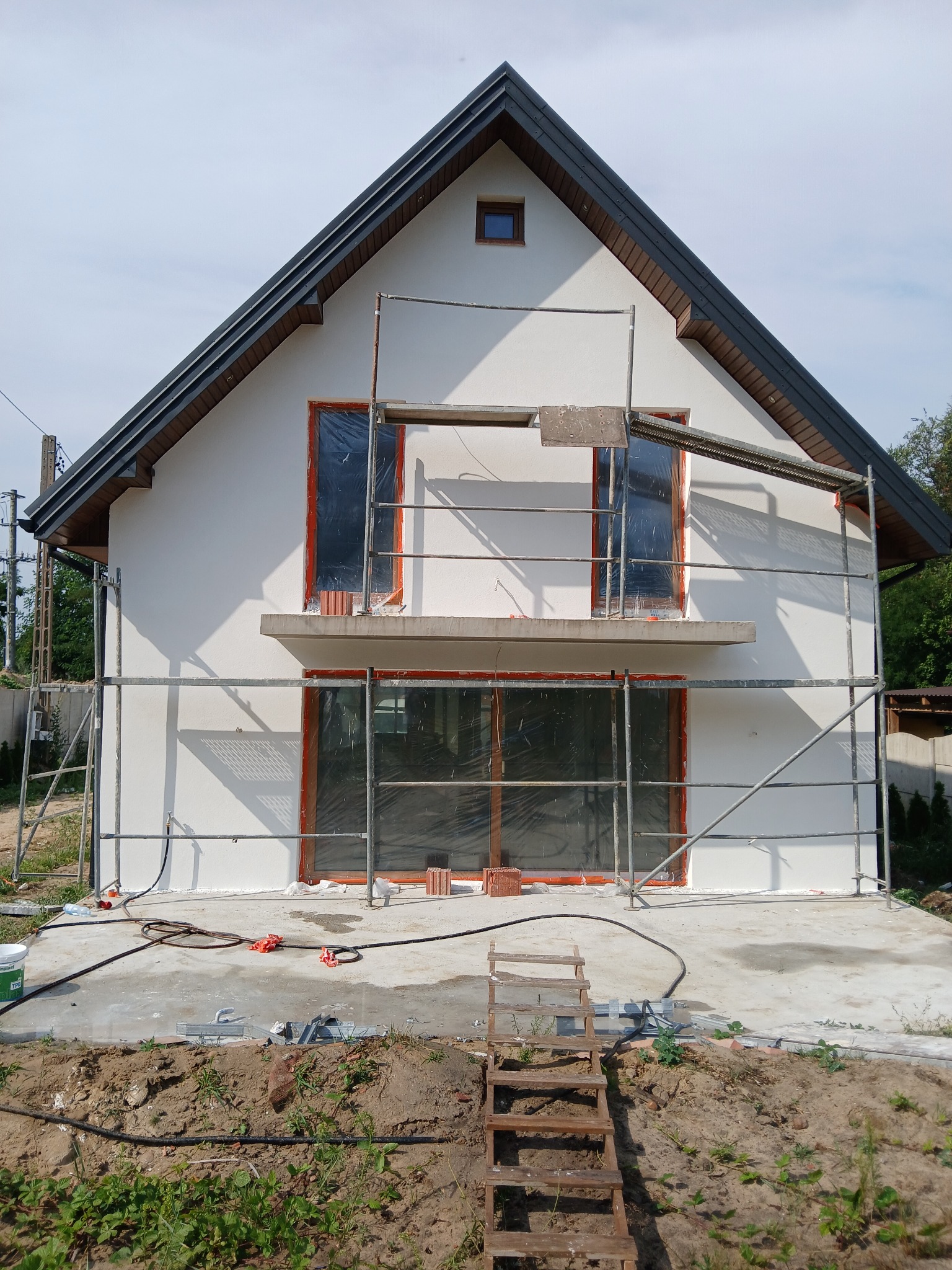 Dom jednorodzinny w trakcie elewacji, z widocznymi rusztowaniami, zabezpieczonymi oknami folią i przygotowanym podłożem wokół budynku.