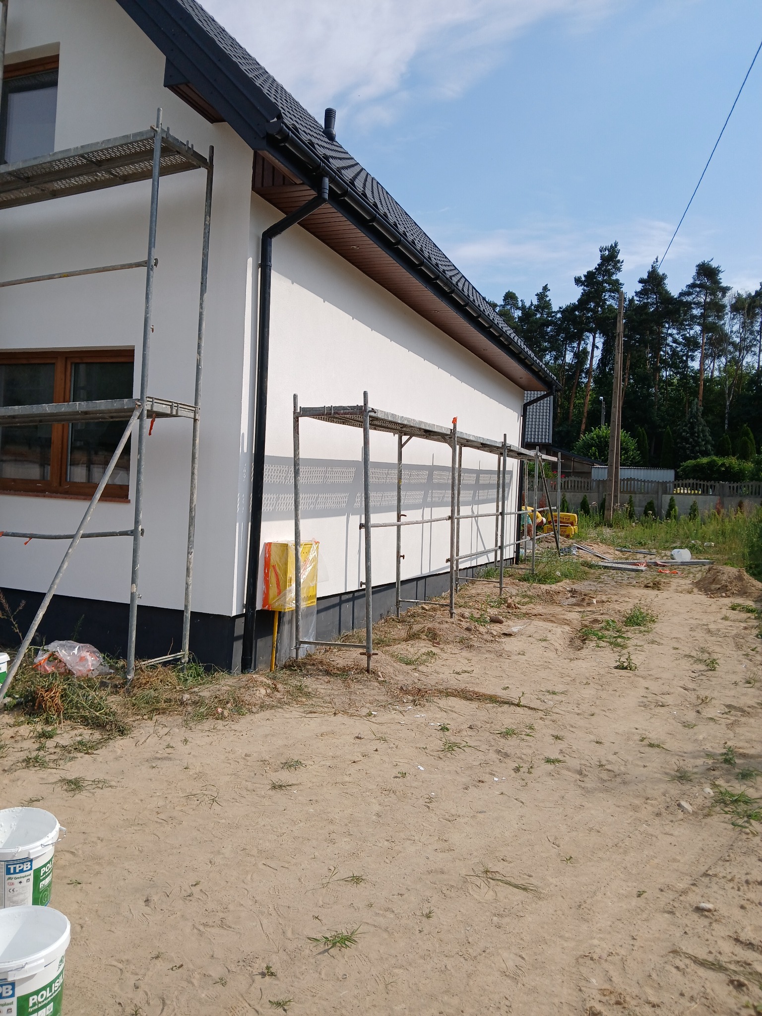 Elewacja domu jednorodzinnego w trakcie prac wykończeniowych, widoczne rusztowania, puszki z farbą i teren wokół w trakcie przygotowania.