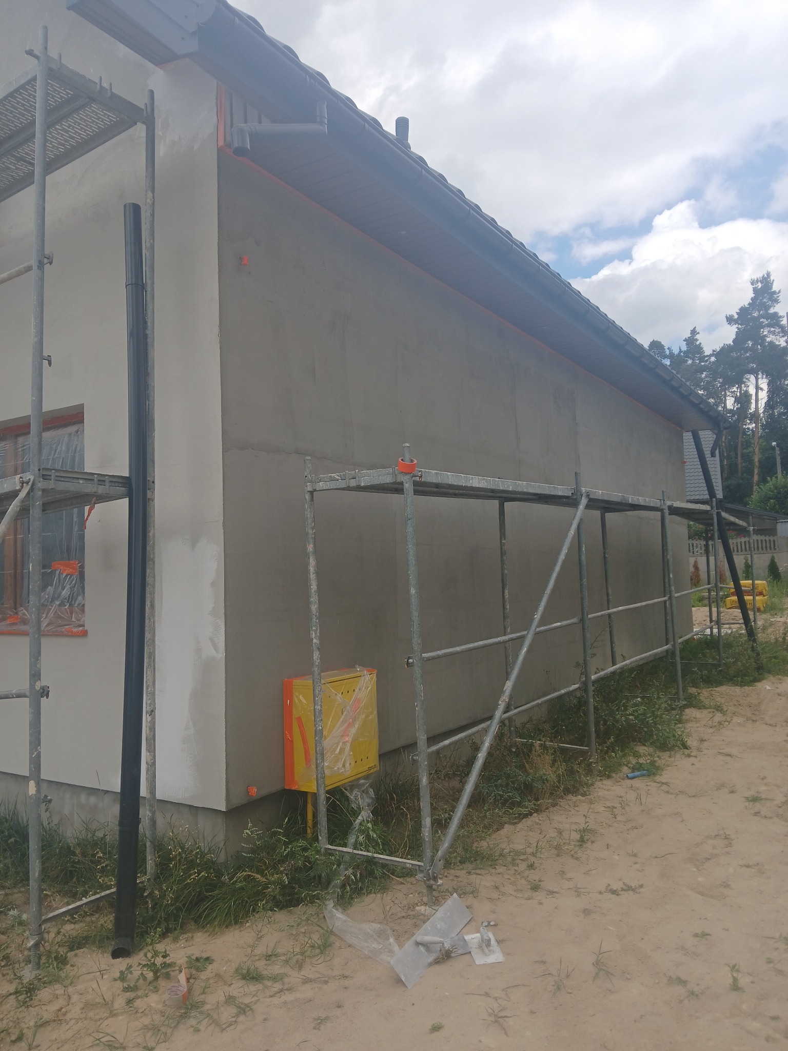 Ściana domu jednorodzinnego w trakcie szpachlowania, widoczne rusztowania, okna zabezpieczone folią, oraz żółta skrzynka gazowa.