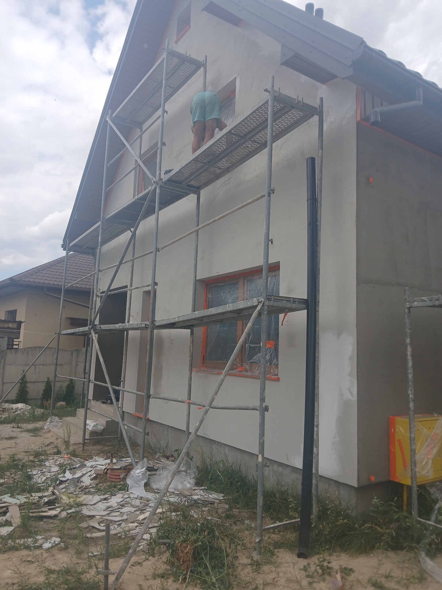 Prace na elewacji domu jednorodzinnego: mężczyzna na rusztowaniu szpachluje ścianę, okna zabezpieczone folią, widoczne ślady po szpachlowaniu. Na ziemi gruz i resztki materiałów budowlanych.