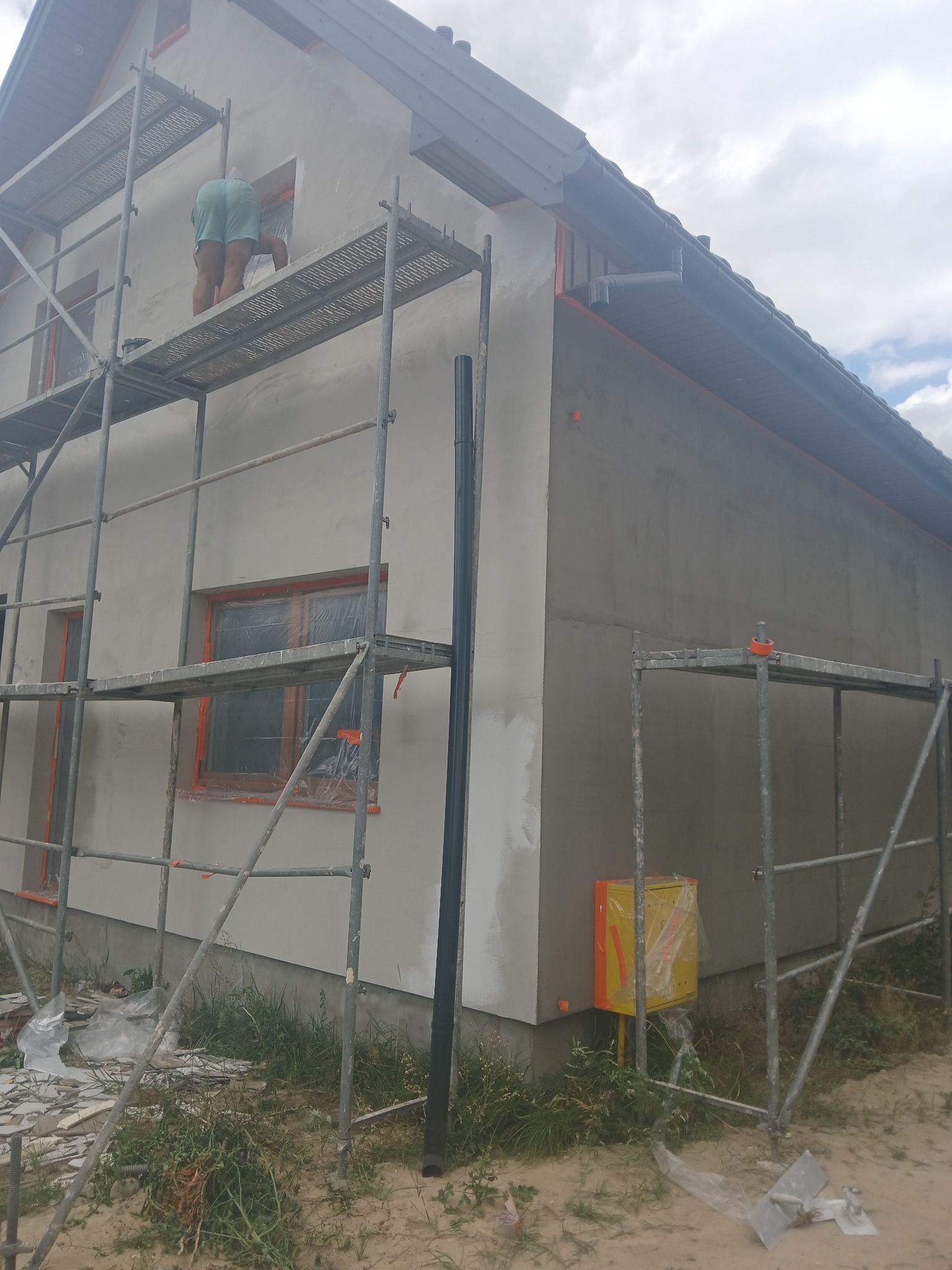 Praca na rusztowaniu: Nakładanie jasnoszarej warstwy tynku na elewację domu jednorodzinnego, okna zabezpieczone folią, widoczna skrzynka gazowa w żółtej obudowie.