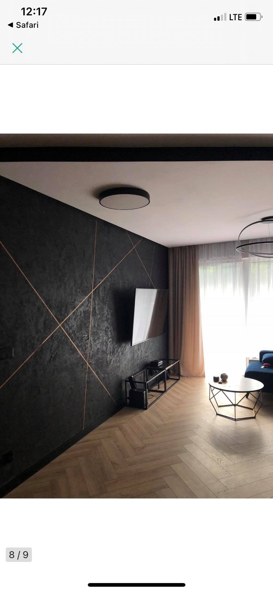 Salon z ciemną ścianą z dekoracyjną fakturą i złotymi liniami, parkiet w jodełkę, telewizor na ścianie, stolik kawowy z metalową podstawą.