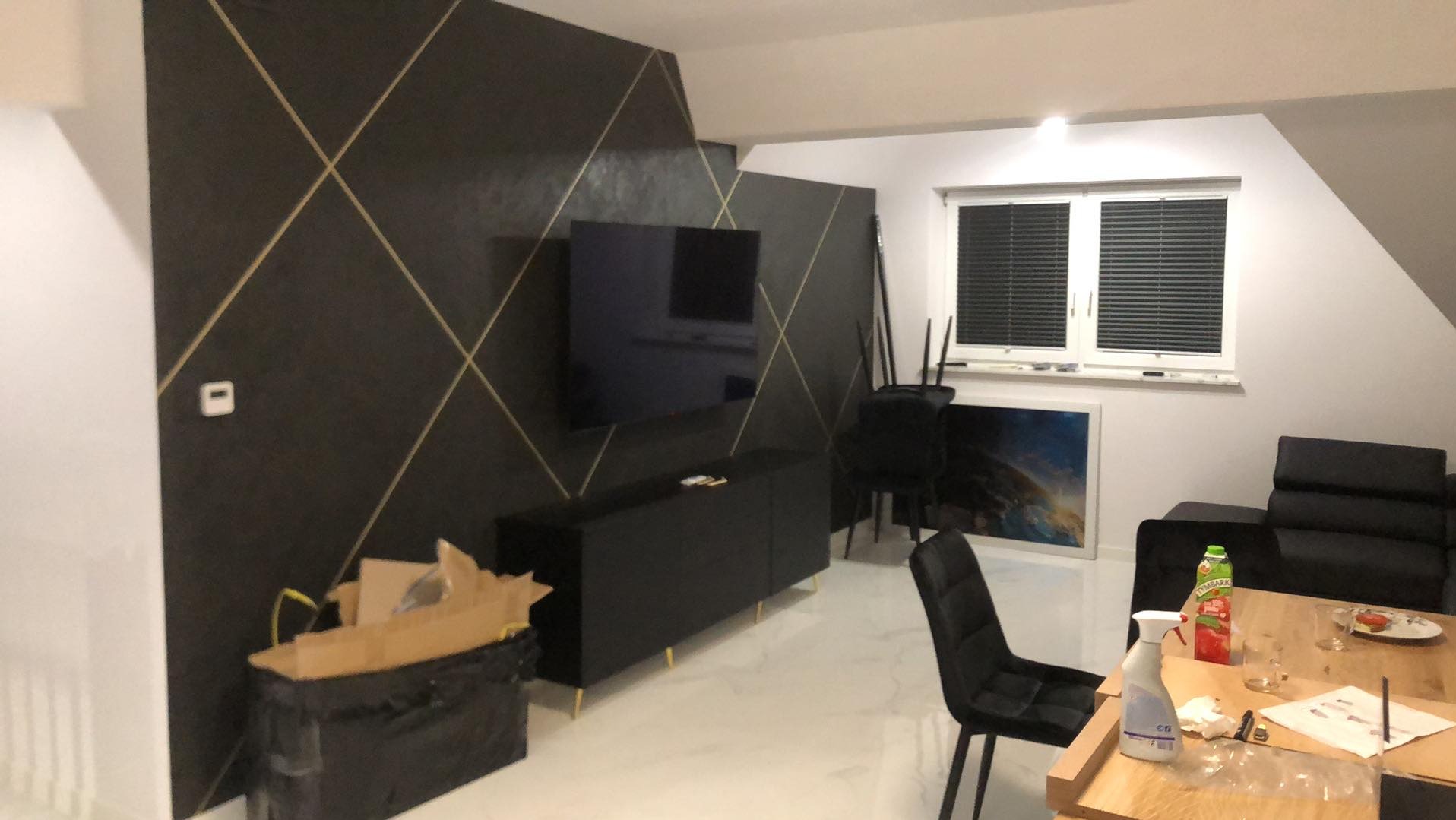 Salon w trakcie wykańczania: czarna ściana z geometrycznym wzorem, czarne meble na złotych nóżkach, biała podłoga, karton z odpadami budowlanymi i nieporządek na stole.