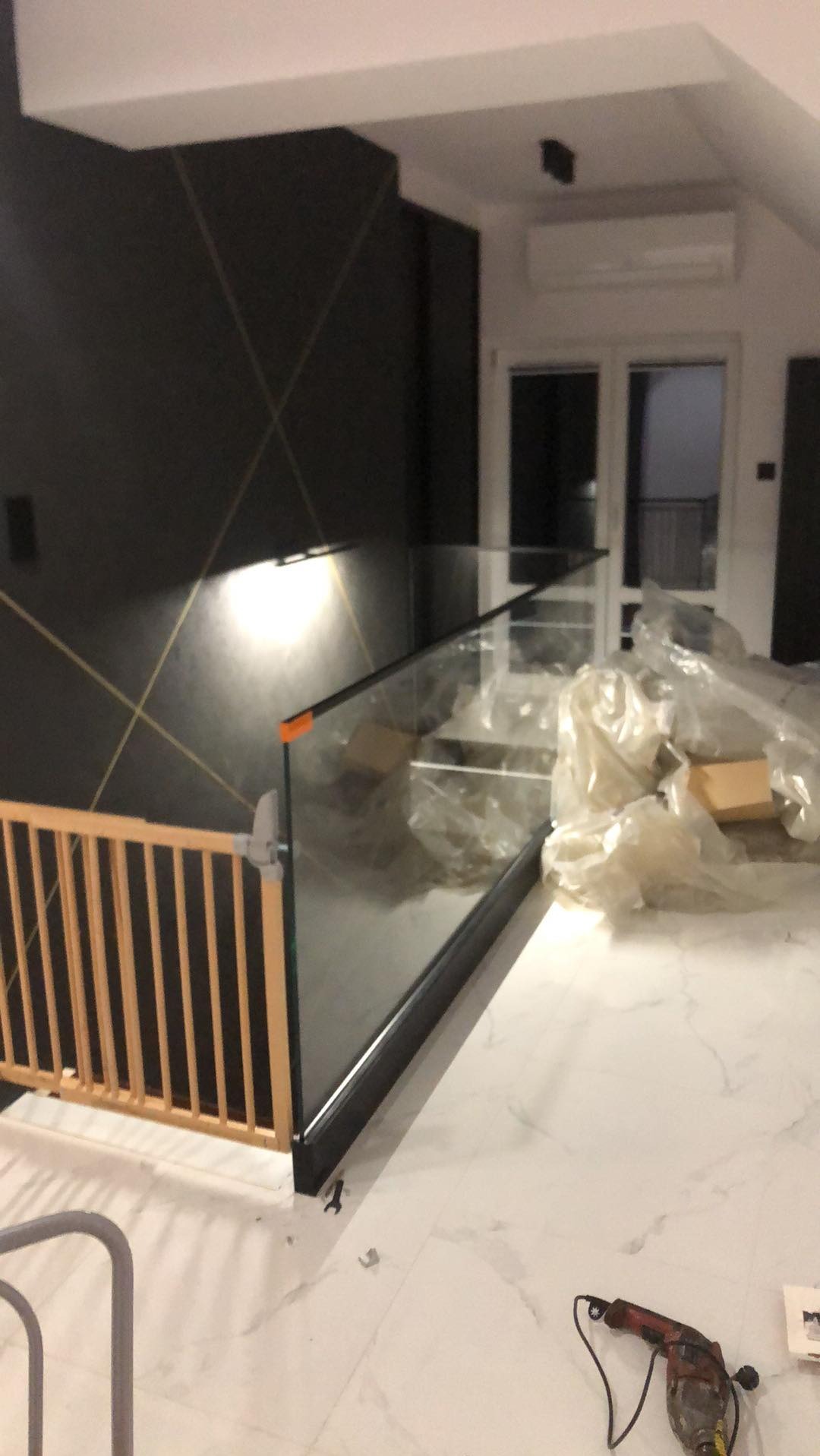 Montaż szklanej balustrady na schodach z zabezpieczeniem dla dzieci, widoczny wiertarko-wkrętak na marmurowej podłodze i fragment czarnej ściany z geometrycznym wzorem.