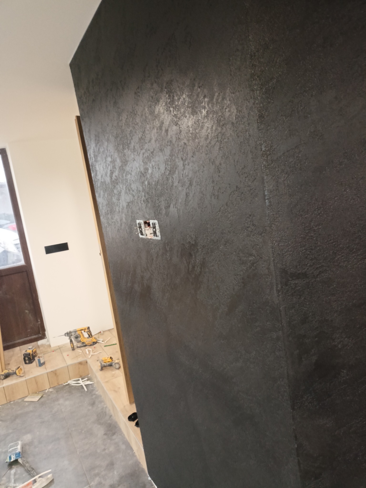 Ściana pokryta ciemnoszarą, strukturalną farbą dekoracyjną, widoczne gniazdko elektryczne, w tle narzędzia budowlane i płytki podłogowe.
