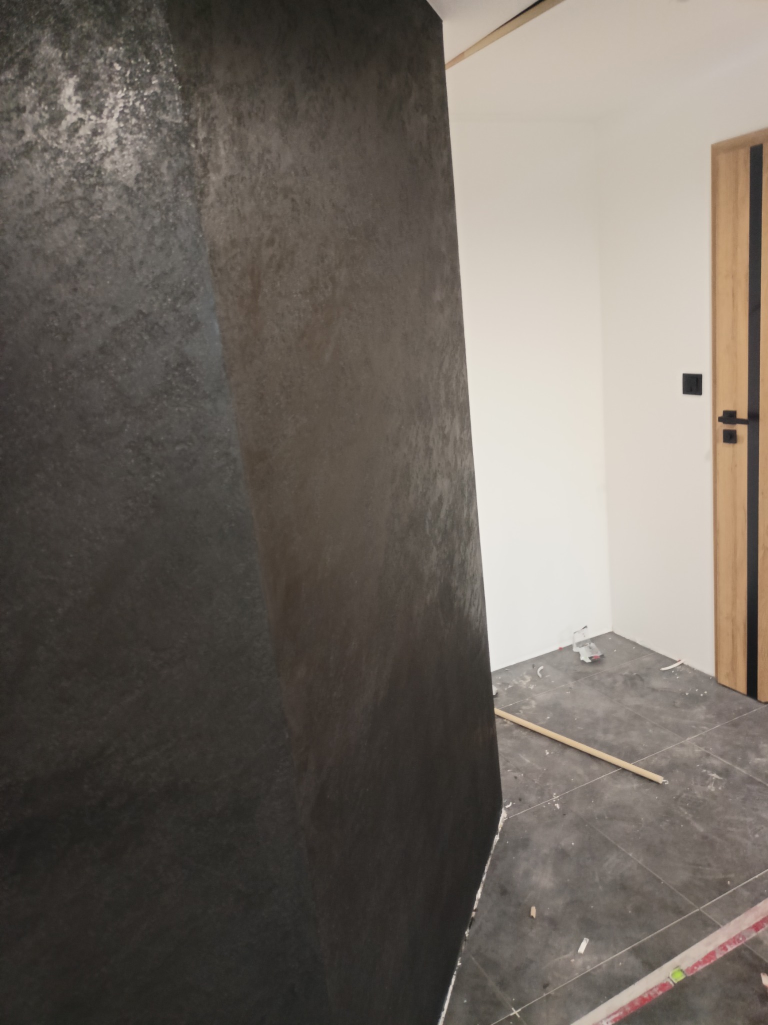 Fragment ściany z dekoracyjną, szarą strukturą przypominającą beton, widoczne narzędzia i zabrudzenia na podłodze z płytek, drzwi wewnętrzne w tle.