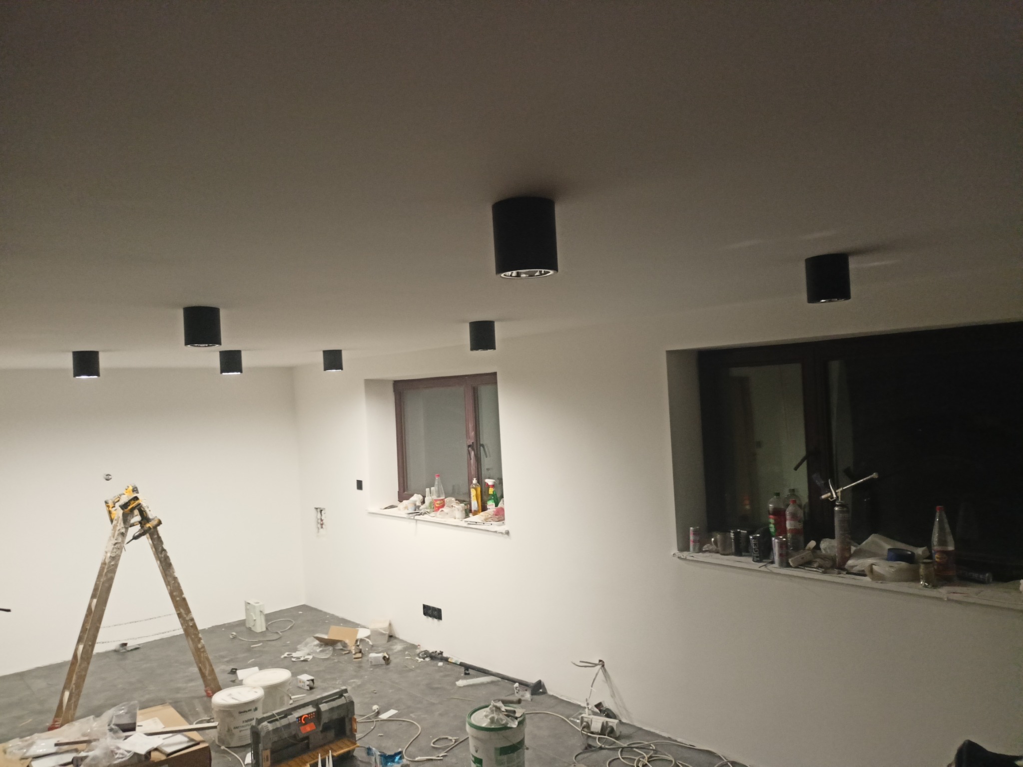 Wnętrze w trakcie remontu: widoczna drabina malarska, puszki z farbą, narzędzia, zamontowane czarne lampy sufitowe, okna z parapetami zastawionymi różnymi przedmiotami.