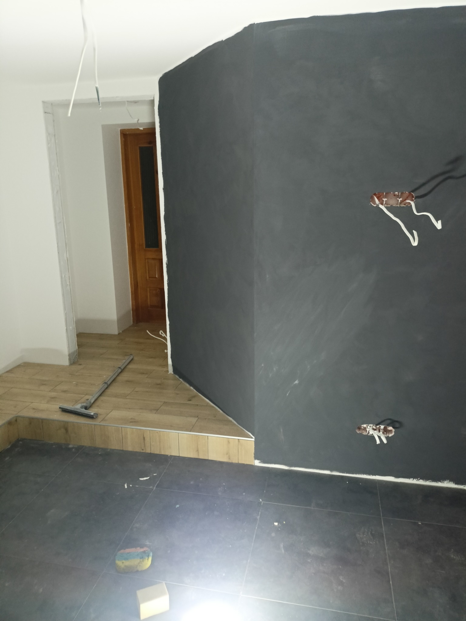 Ciemnoszara ściana z widocznymi puszkami elektrycznymi bez osprzętu, fragment drewnianej podłogi z listwą przypodłogową i narzędziem do czyszczenia podłóg, ciemne płytki podłogowe z gąbką i kostką,...