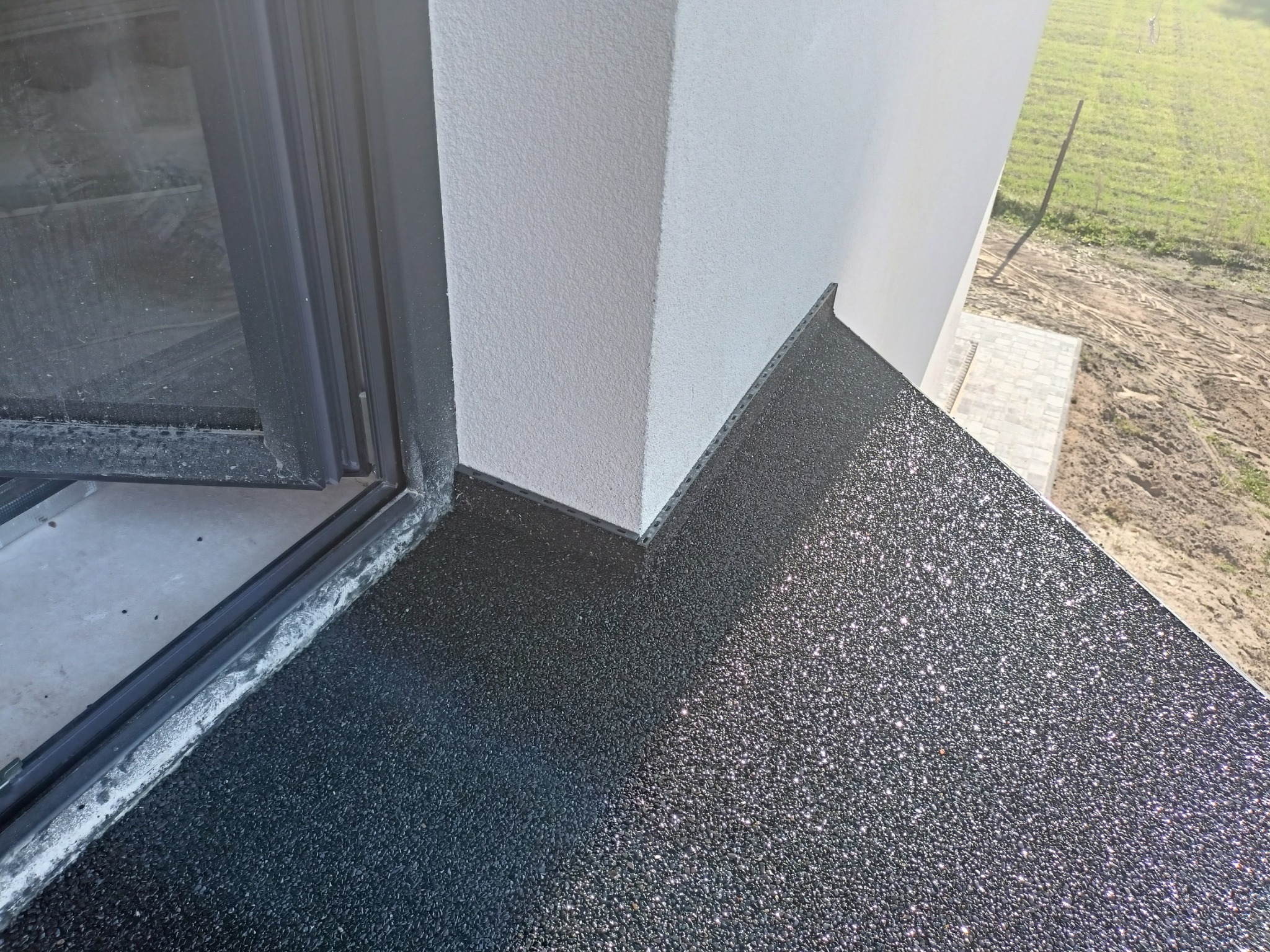 Czarna, błyszcząca powierzchnia balkonu z żywicy epoksydowej, widok z góry, z fragmentem okna balkonowego i elewacji budynku.
