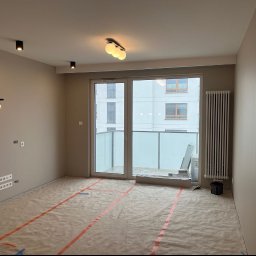 NEW-HOUSEBUD SPÓŁKA Z OGRANICZONĄ ODPOWIEDZIALNOŚCIĄ - Przygotowane do malowania wnętrze mieszkania z zabezpieczoną podłogą, widoczne puszki elektryczne i grzejnik w tle, nowoczesne oświetlenie sufitowe.