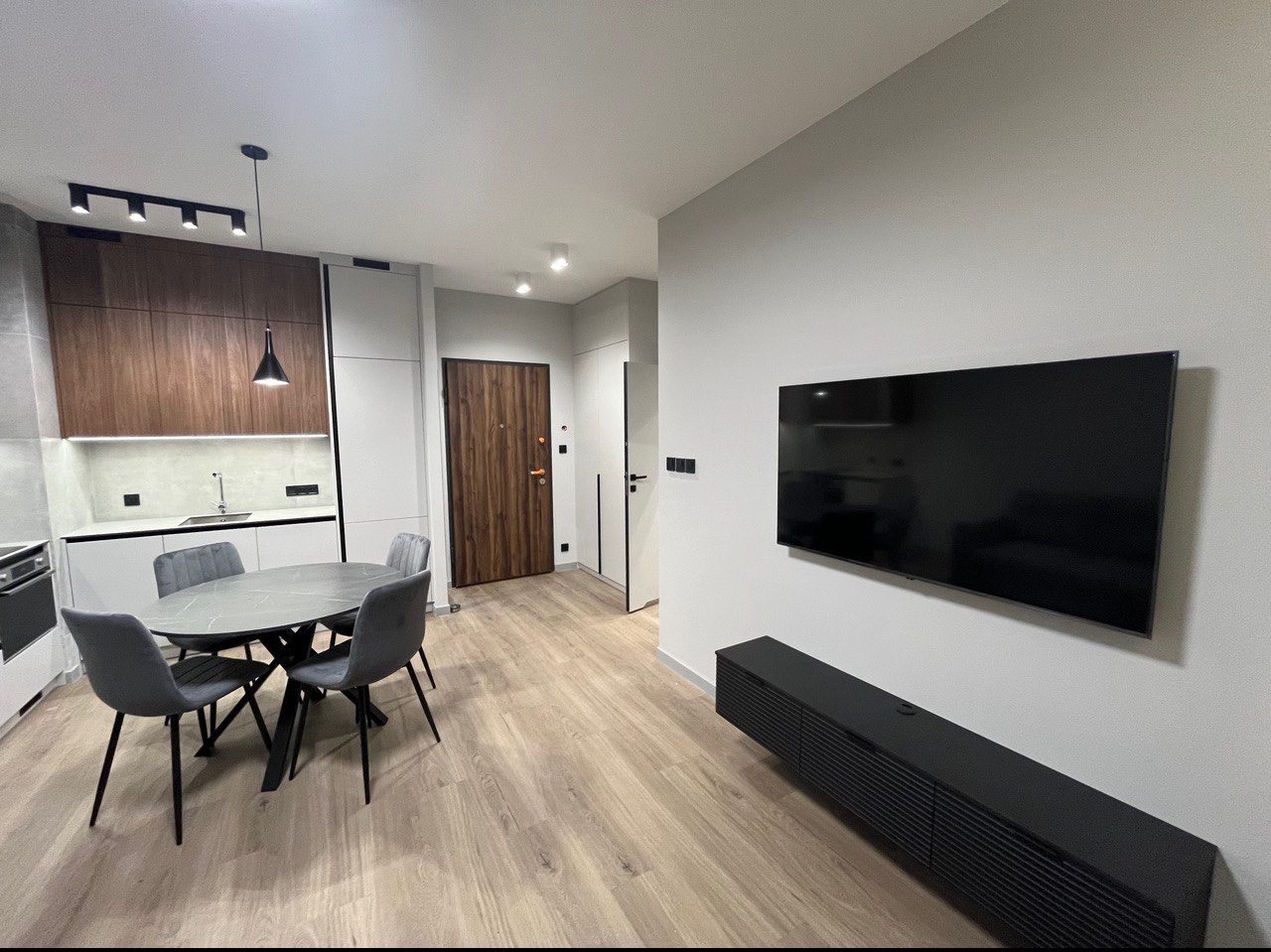 Nowoczesne wnętrze mieszkania z drewnianą podłogą, szarą okrągłą jadalnią z czterema krzesłami, zabudową kuchenną z drewnianymi i białymi frontami oraz czarnym telewizorem na ścianie.