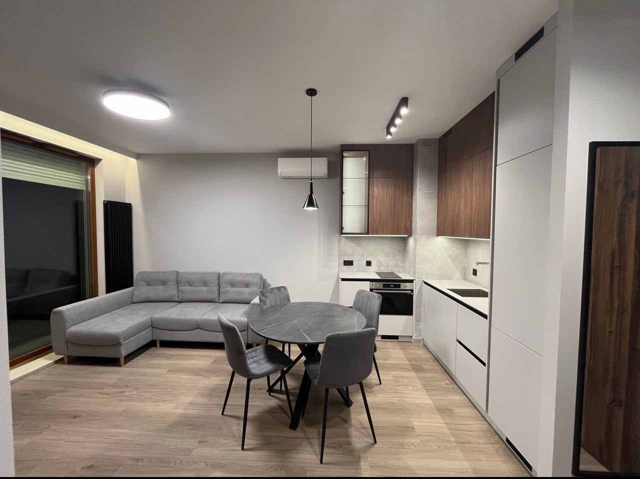 Jasne, przestronne wnętrze mieszkania z szarą sofą narożną, okrągłym stołem z czarnymi nogami i krzesłami w odcieniach szarości, oraz nowoczesną kuchnią z białymi szafkami i drewnianymi frontami.