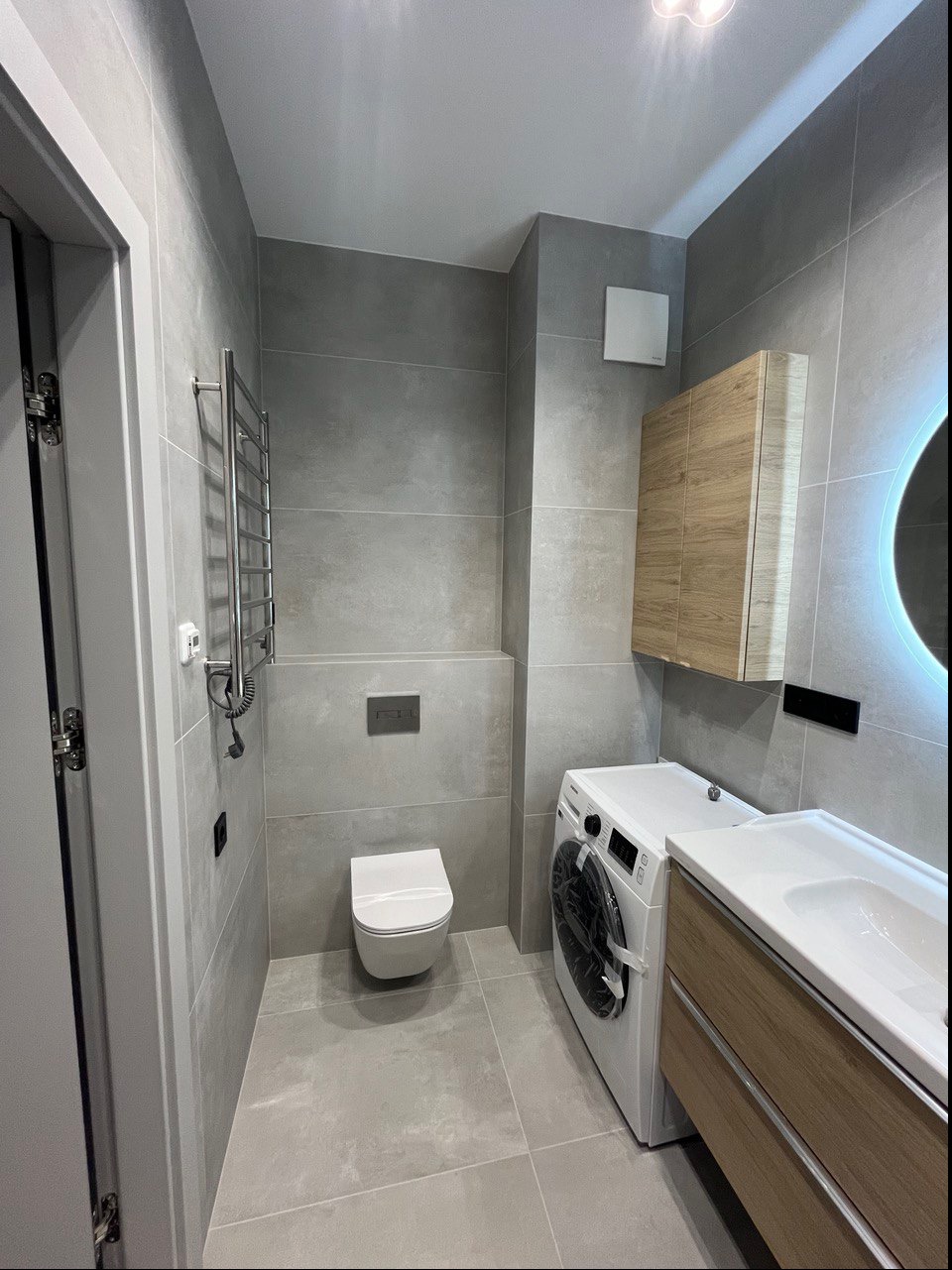 Wykończona łazienka w szarościach z podświetlanym lustrem, wiszącą toaletą, grzejnikiem drabinkowym i pralką w folii ochronnej.