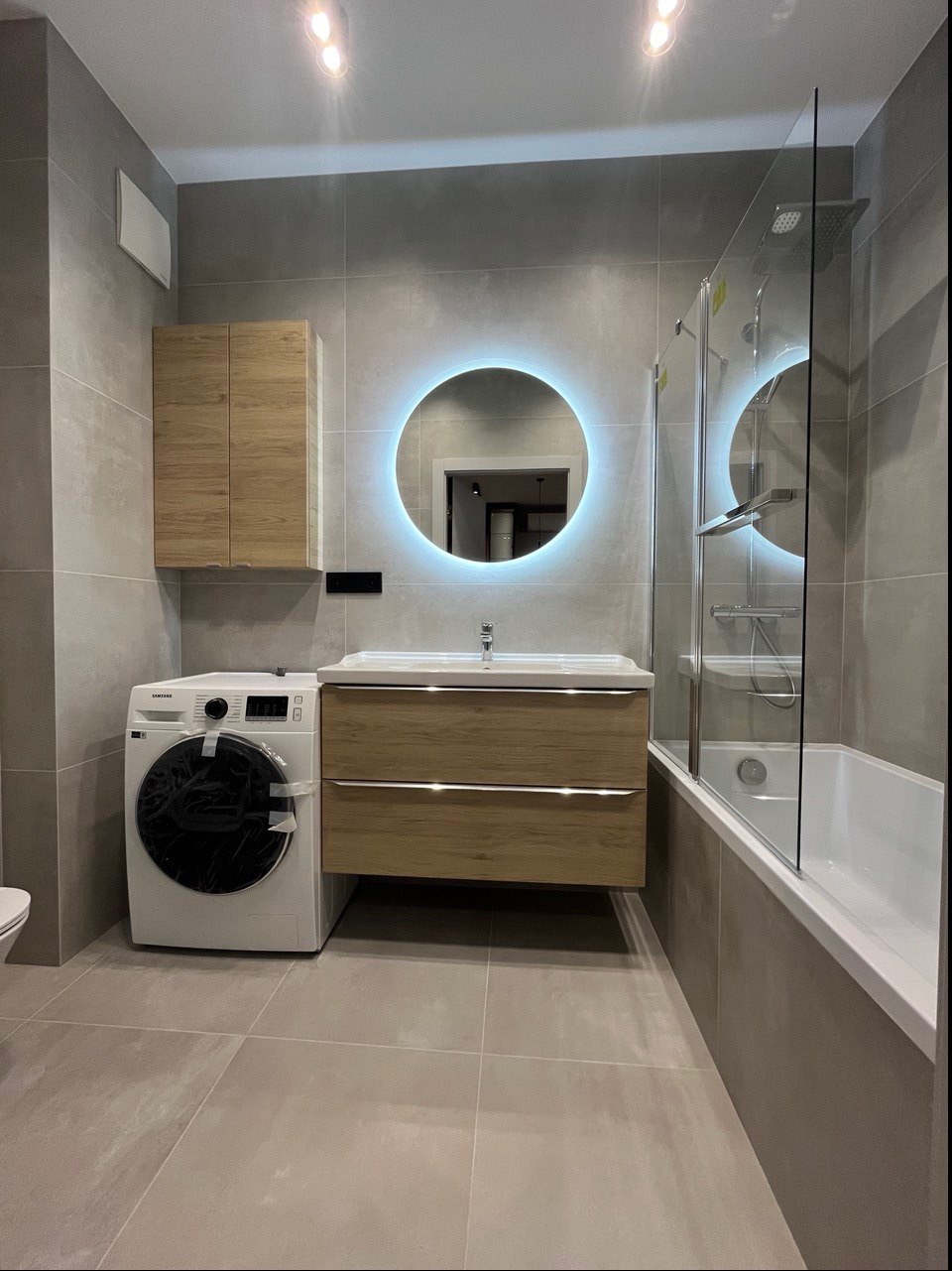 Nowoczesna łazienka w szarościach z okrągłym lustrem podświetlanym LED, drewnianą szafką pod umywalką i pralką Samsung z folią ochronną.