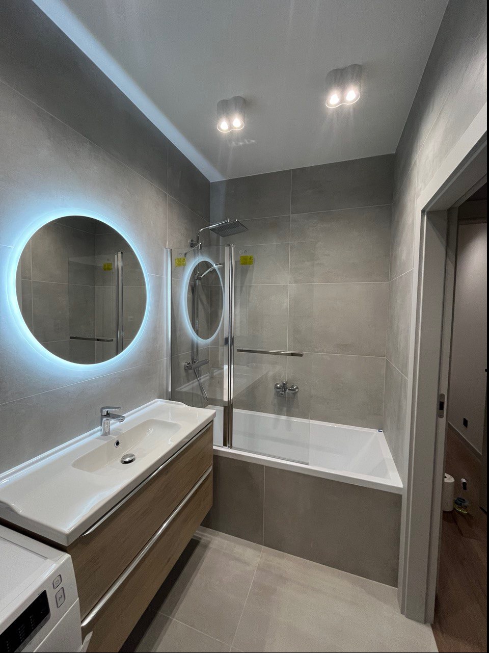 Nowoczesna łazienka w szarościach z podświetlanym lustrem, drewnianą szafką pod umywalką i kabiną prysznicową nad wanną.