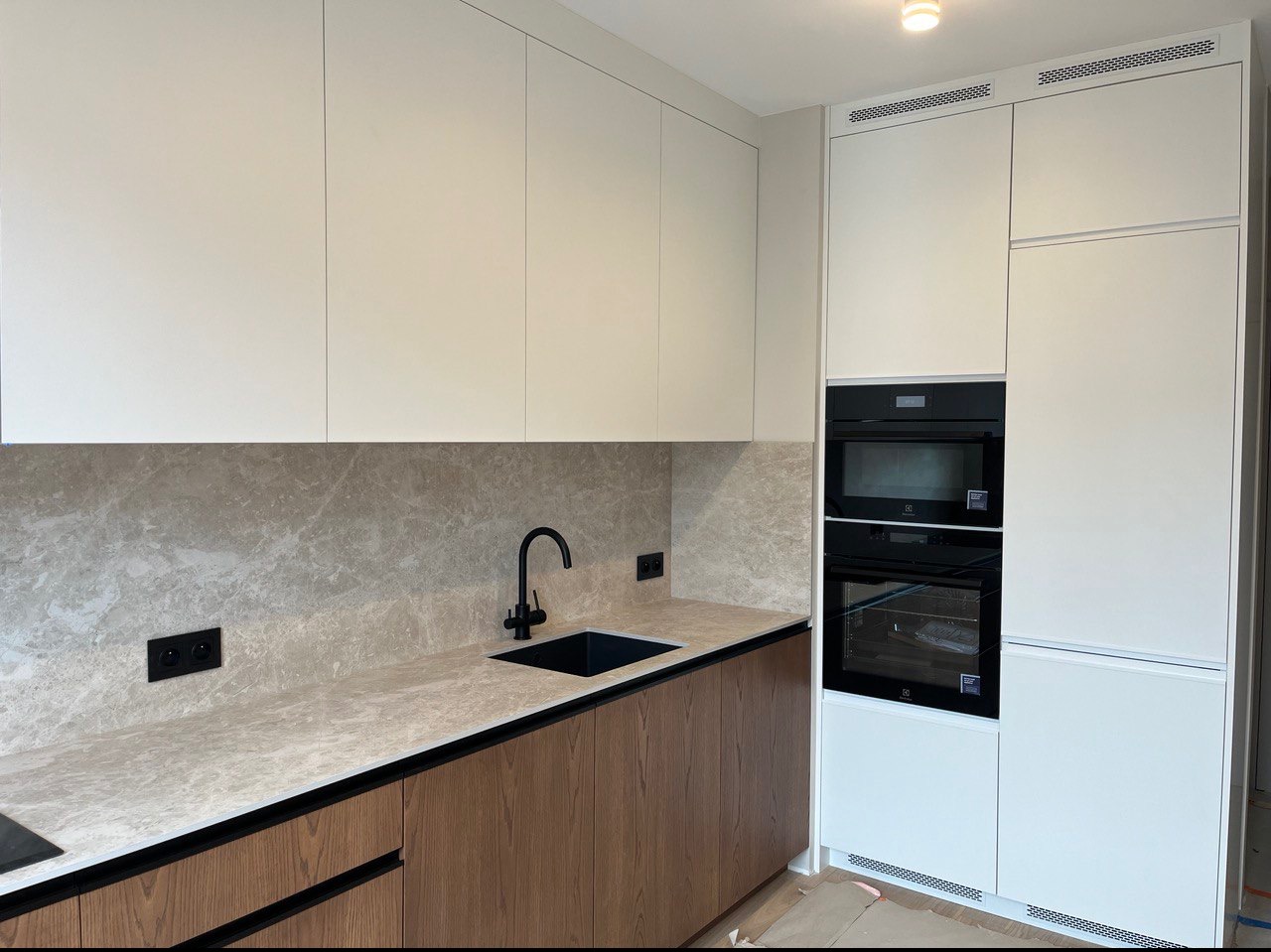 Nowoczesna kuchnia z białymi górnymi szafkami, drewnianymi dolnymi szafkami z czarnymi uchwytami, marmurowym blatem, czarną baterią i zlewem oraz zabudowanymi piekarnikiem i mikrofalówką Electrolux.