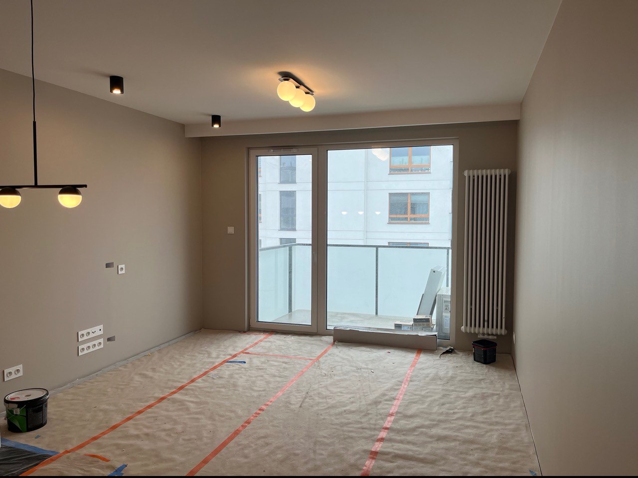 Przygotowane do malowania wnętrze mieszkania z zabezpieczoną podłogą, widoczne puszki elektryczne i grzejnik w tle, nowoczesne oświetlenie sufitowe.