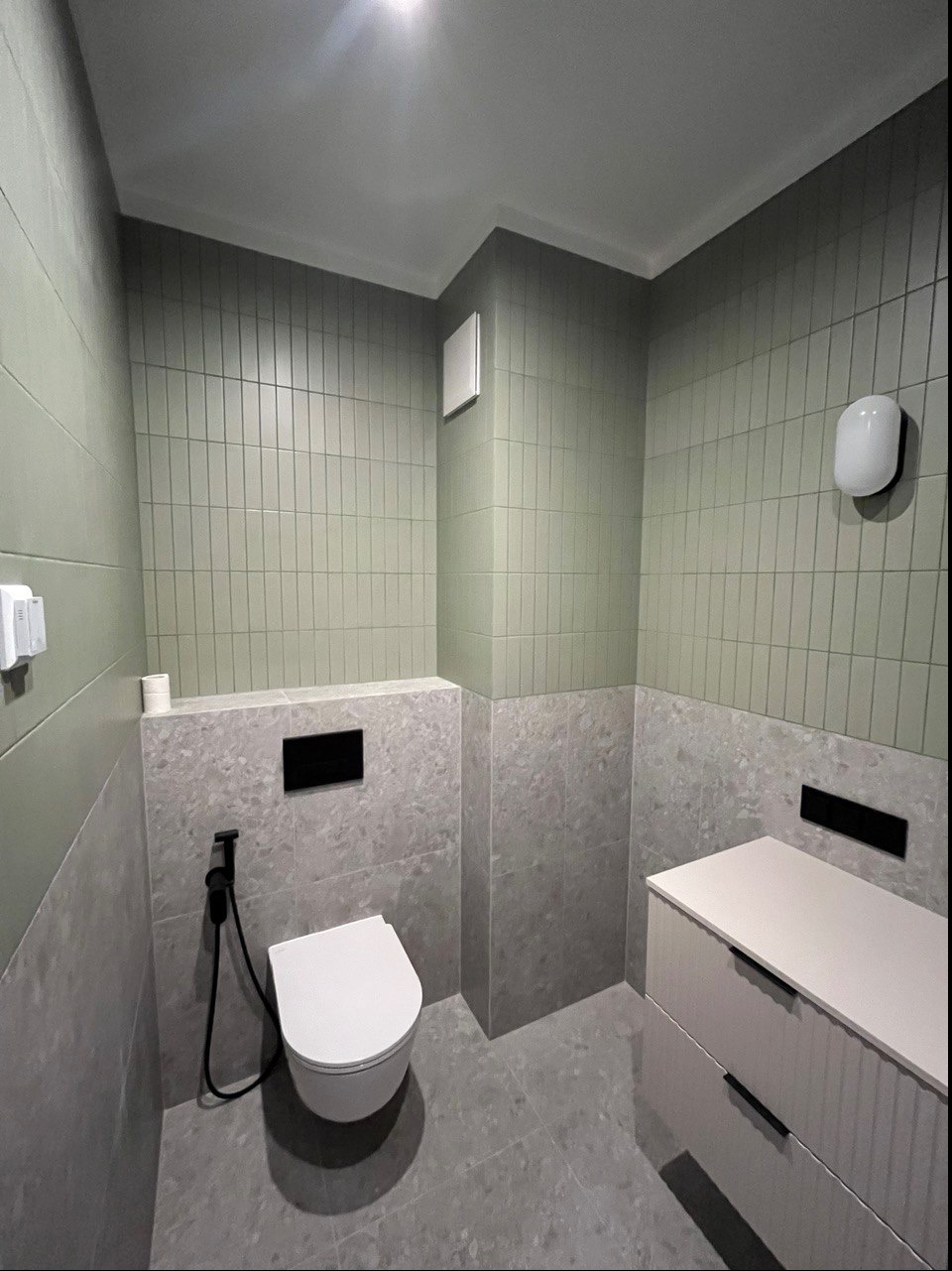 Nowoczesna łazienka z pionowymi, oliwkowymi płytkami na ścianach, szarą podłogą i zabudową podtynkową WC z bidetką, minimalistyczna szafka z szufladami i czarnymi uchwytami.