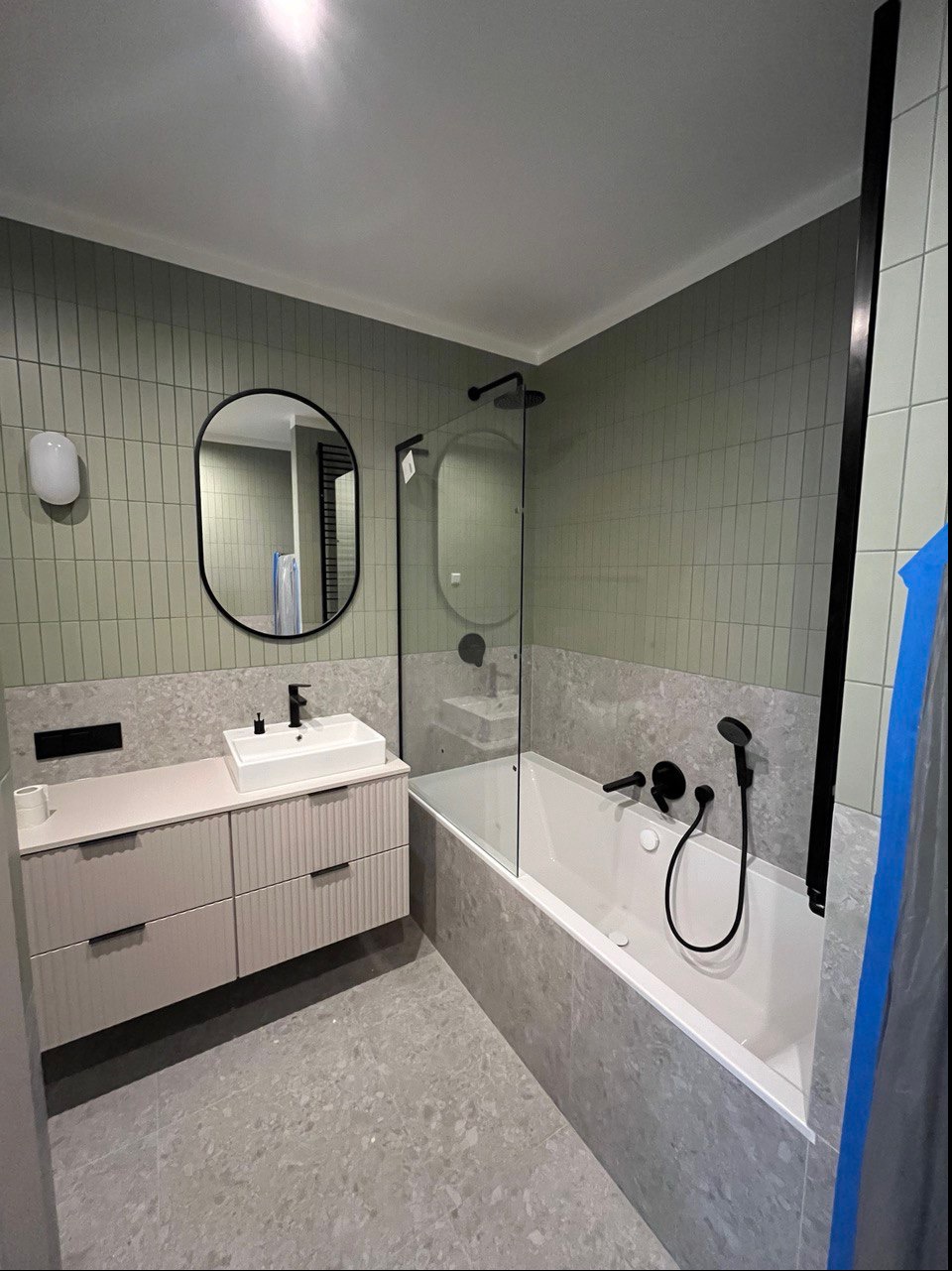 Nowoczesna łazienka w odcieniach szarości i zieleni z minimalistyczną umywalką, owalnym lustrem w czarnej ramie i wanną z czarną armaturą oraz szklanym parawanem.