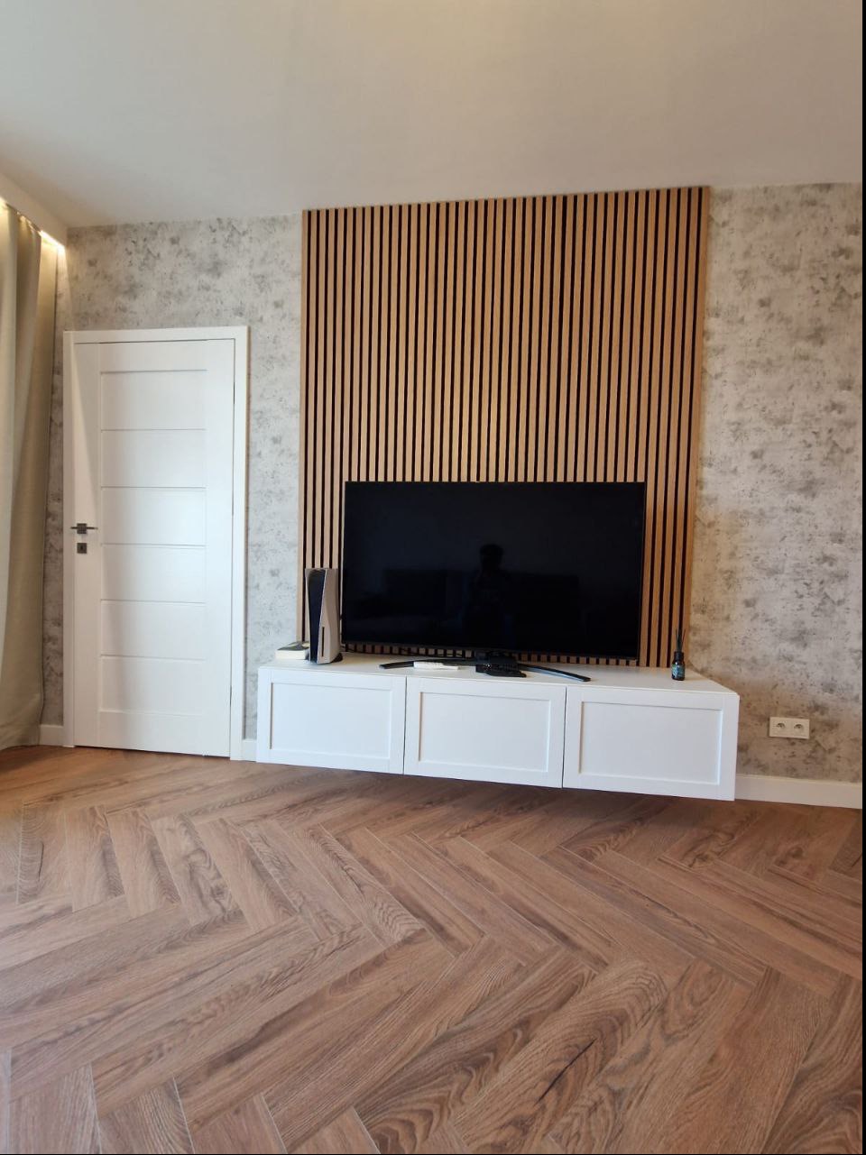 Salon z drewnianą ścianą z lameli, telewizorem na białej szafce RTV, parkietem ułożonym w jodełkę i białą ścianą z efektem betonu.