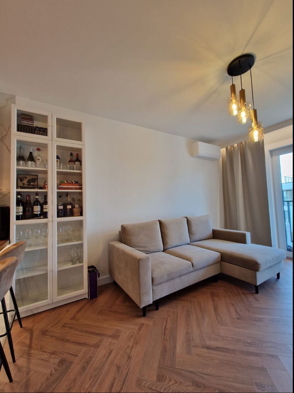 Jasny salon z beżową sofą narożną, drewnianą podłogą ułożoną w jodełkę, regałem z białymi ramami wypełnionym szkłem i butelkami oraz nowoczesną lampą wiszącą z trzema szklanymi kloszami.