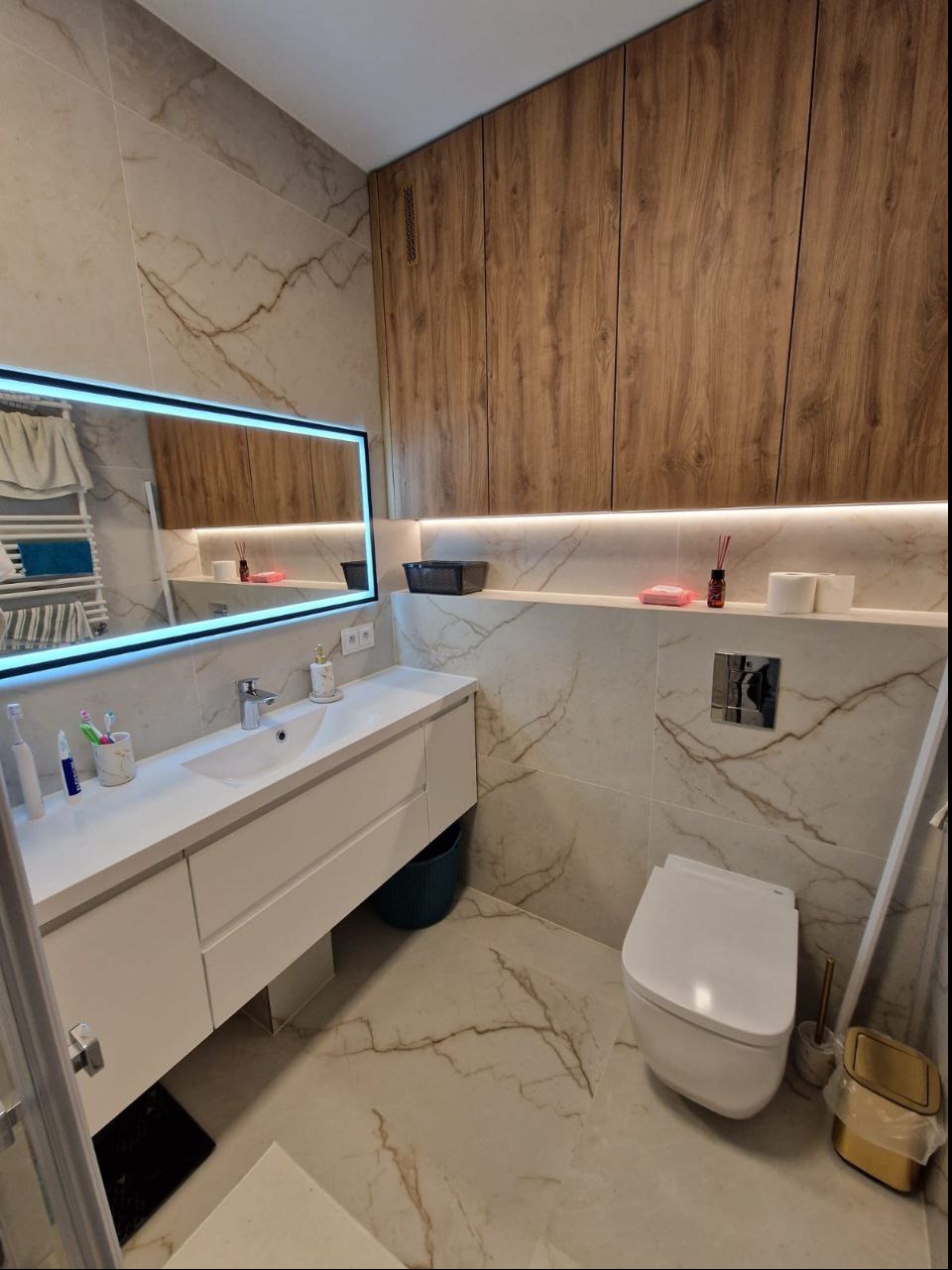 Nowoczesna łazienka z marmurowymi płytkami, podświetlanym lustrem LED, białą szafką umywalkową i drewnianymi szafkami wiszącymi, minimalistyczna toaleta, widoczne akcesoria łazienkowe.