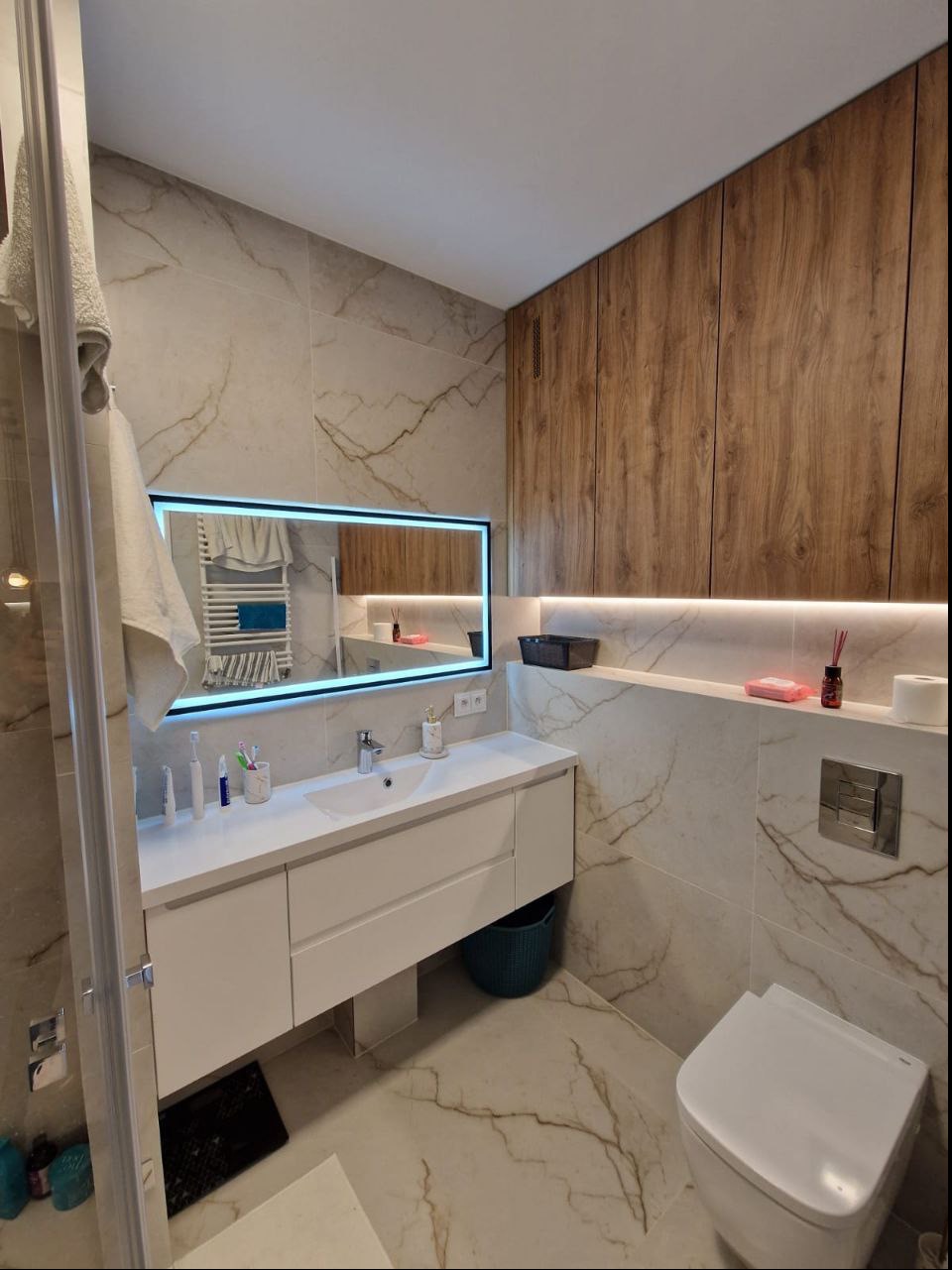 Nowoczesna łazienka z marmurowymi płytkami, podświetlanym lustrem i drewnianą szafką wiszącą, widok na umywalkę z białym blatem i toaletę.
