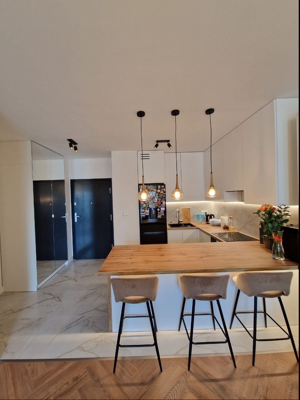 Nowoczesna kuchnia z drewnianym blatem wyspowym, trzema hokerami z beżowymi siedziskami i wiszącymi lampami z dekoracyjnymi żarówkami, podświetlany marmurowy backsplash.